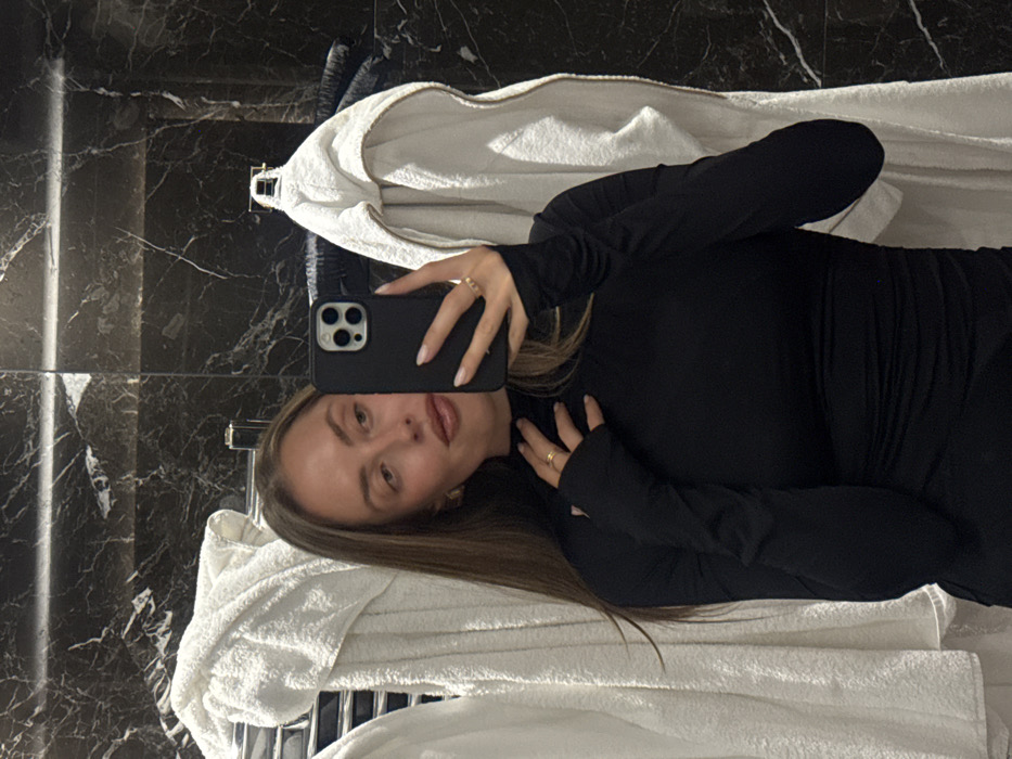 A woman takes a mirror selfie. She is wearing a black long-sleeved shirt and gold jewelry. Her hair is brown and her nails are painted light pink. She holds a black smartphone in her right hand. A white towel lies in the background. The bathroom has black and white marble tiling and chrome hardware.