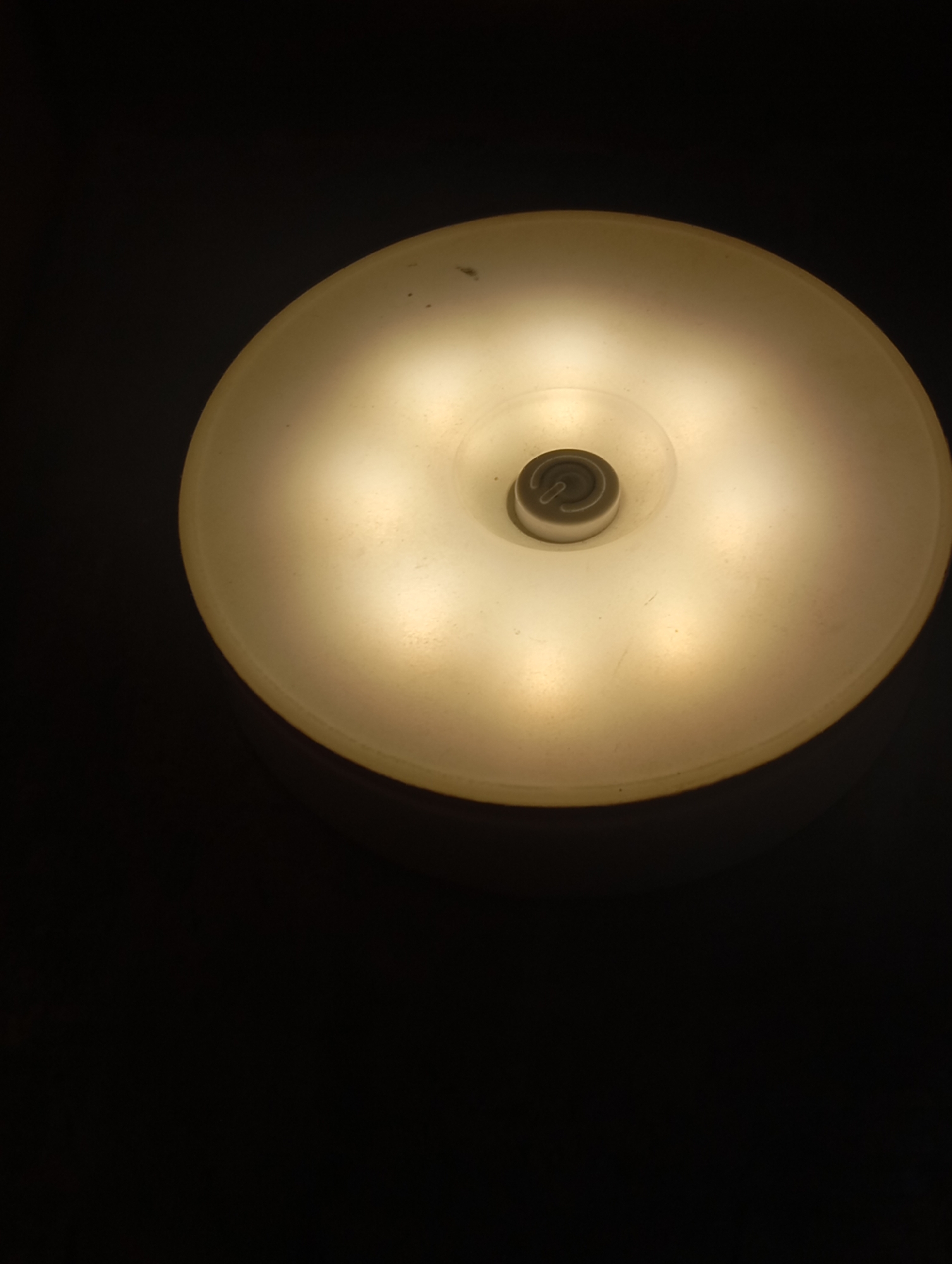 A dimly lit, close-up shot focuses on a round, translucent light fixture against a black background. The fixture emits a warm, yellow glow, illuminating the surface of the light. In the center of the fixture, a small, circular silver component with a spiral design is visible. The light appears to be a ceiling or overhead light. The setting is indoors, in a dark environment, with no specific city or location identifiable.