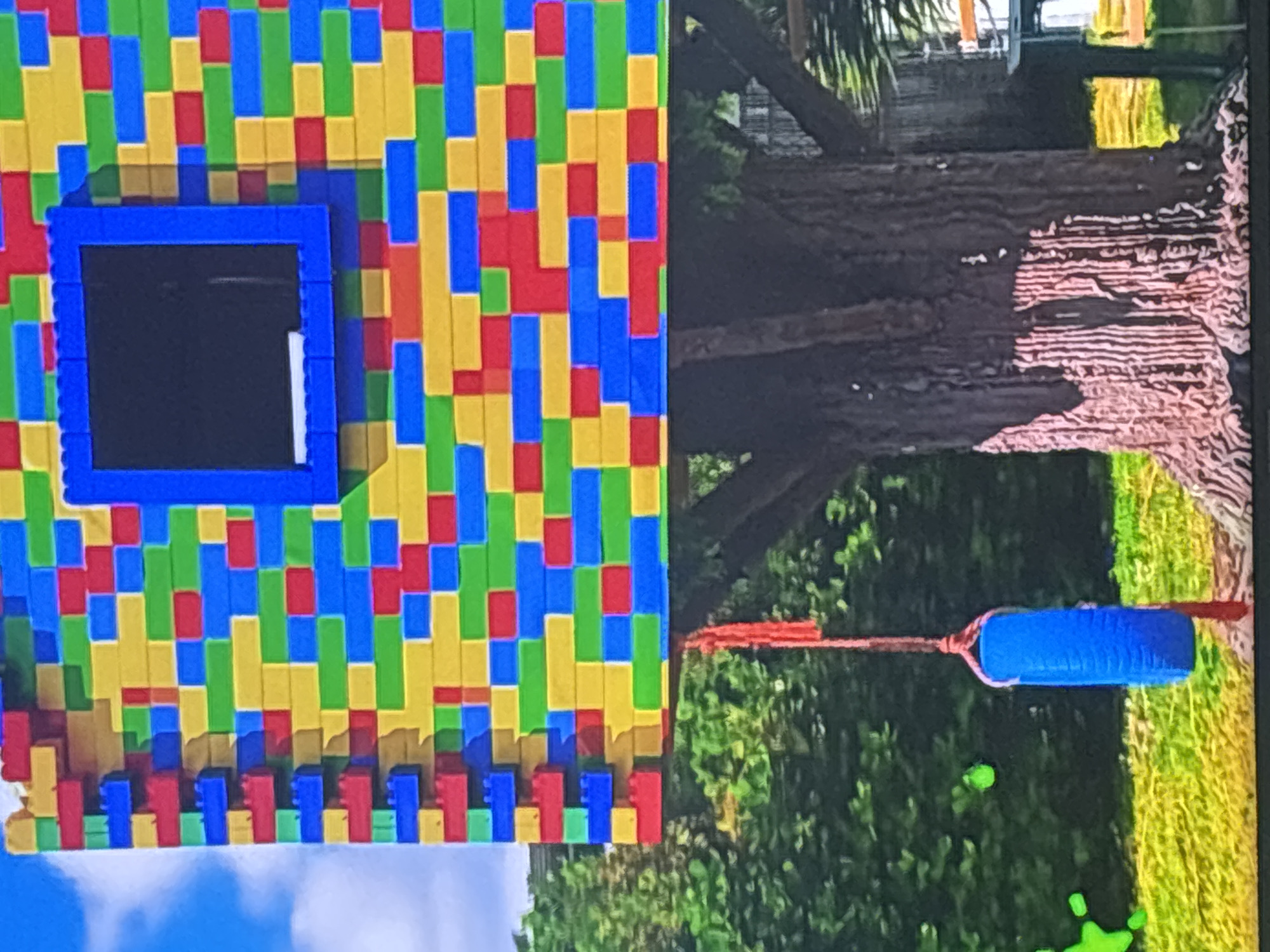 The image shows a whimsical treehouse constructed from large, colorful building blocks, resembling giant LEGO bricks. The treehouse is mounted on a large tree, with a blue tire swing hanging from a red rope. The tree is situated in a grassy area with lush green foliage in the background. The sky is partly cloudy, with patches of blue visible. The treehouse has a dark square opening that appears to be a window. The overall scene suggests an outdoor recreational space, likely a backyard or a park. There is no information in the image to suggest it is located in Dubai, United Arab Emirates, or any other specific city.