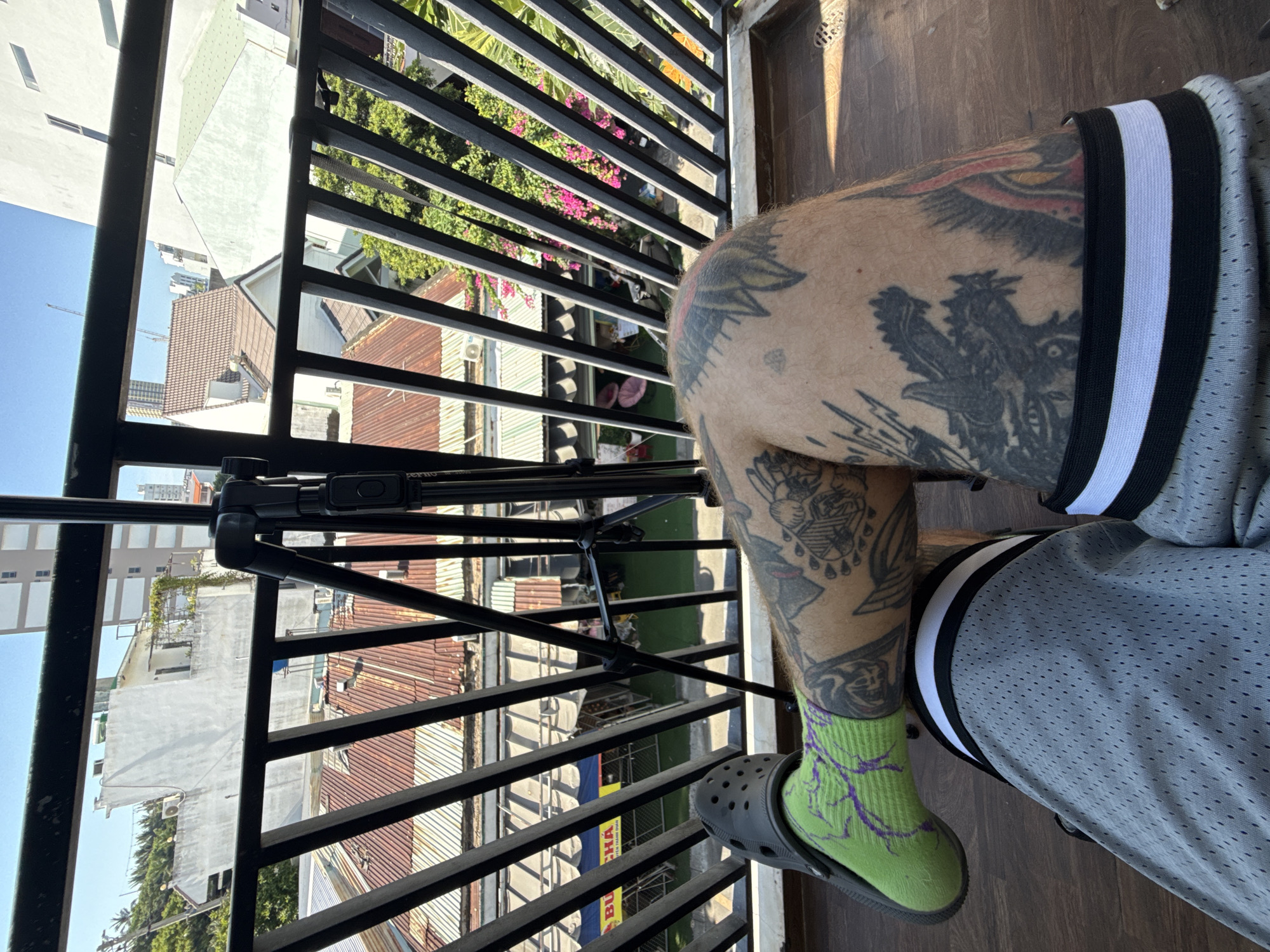 The image shows a partial view of a person with heavily tattooed legs sitting on a balcony in Đà Nẵng, Vietnam. The person is wearing grey athletic shorts with black and white trim, a bright green sock with purple designs, and a grey Croc. The balcony railing is black metal. A tripod sits in front of the railing, and the view beyond the railing shows closely packed buildings with rusty roofs and some greenery, typical of urban Đà Nẵng. There is a white building in the background, and part of the clear blue sky is also visible.