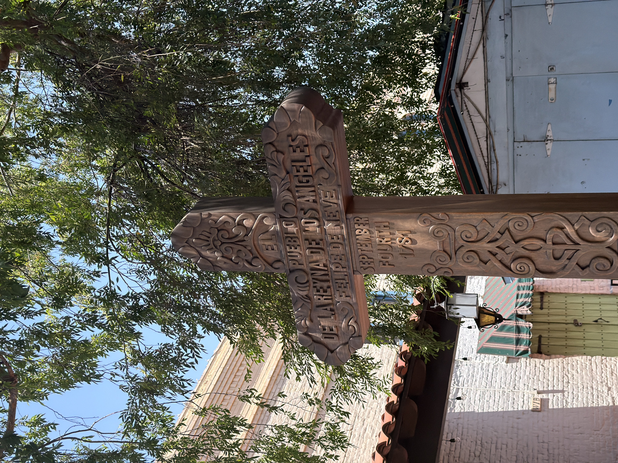 The image depicts a brown wooden sign in the shape of a cross, placed in a location within the city of Los Angeles, United States of America. The sign features carved text, partially obscured by tree branches. The visible text reads "EL PUEBLO DE LOS ANGELES DE LA REYNA DE FELIPE DE NEVE SEPTEMBER FOURTH 1781." The sign is positioned against a backdrop of buildings with varying architectural styles, including a structure with a tiled roof and white walls, and another with light blue panels. The presence of a street lamp indicates an urban environment. The scene appears to be outdoors, captured during daylight hours, with sunlight filtering through the foliage.