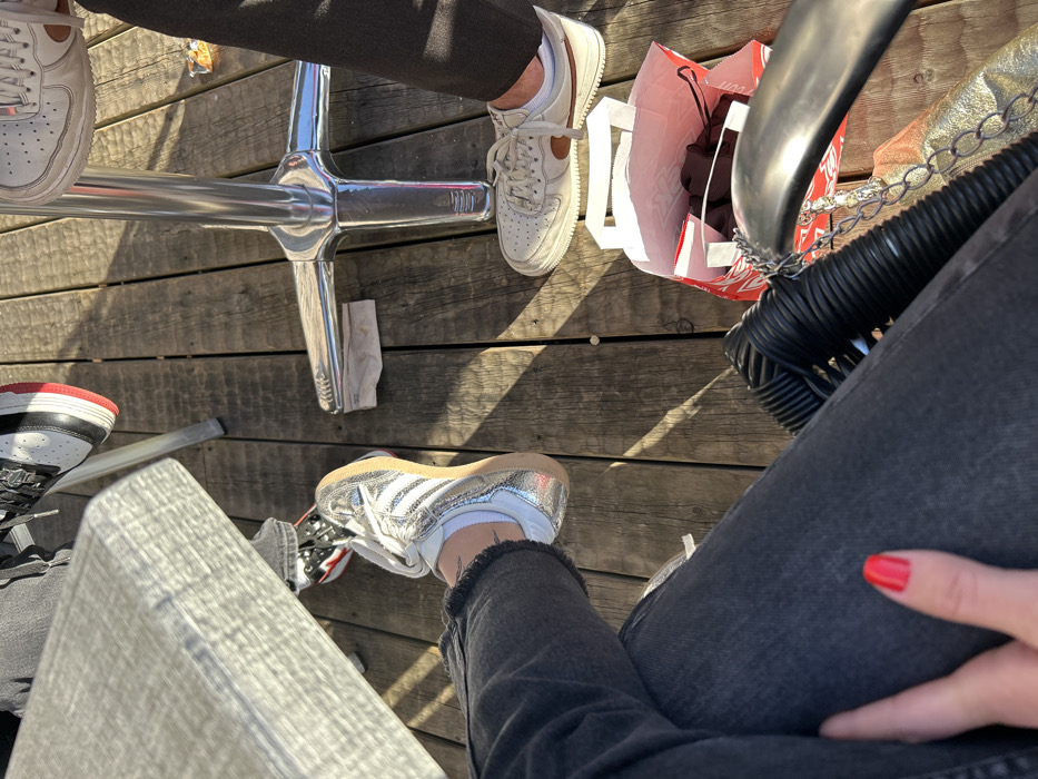 The image captures a high-angle view looking down at the feet and legs of at least three individuals seated around a table on a wooden outdoor deck. Strong sunlight casts distinct shadows across the wooden planks, suggesting it is a clear day, likely late morning or early afternoon.
In the upper central part of the frame, a shiny, chrome-plated table base with four radiating legs is prominent. To the left of this base, a person's leg clad in dark pants is visible, along with a white sneaker featuring tan or brown accents, possibly a Nike Air Force 1 style. A white paper napkin or tissue lies on the wooden floor beneath the table base.
To the right of the table base, another person's foot is visible, wearing a similar white sneaker with tan accents. Next to this foot is a red shopping bag with white handles, appearing to hold dark fabric, possibly clothing. Part of a black wicker chair or stool is seen next to the bag, with a metallic chain (possibly from a handbag) draped over it. A shimmering, metallic handbag is partially visible behind the wicker chair.
In the lower left quadrant, a leg wearing light grey jeans and a black and white sneaker with red accents (resembling a Jordan 1) is present. Further to the right, in the foreground, another person's legs are visible, wearing dark grey or black distressed jeans with a frayed hem above white socks. This person is wearing striking silver or metallic sneakers, possibly an Adidas Samba style. A hand with bright red nail polish rests on the thigh of the person in the foreground, near the bottom right corner of the image.
The overall scene suggests a casual outdoor gathering, possibly at a cafe or restaurant with deck seating. No discernible text is visible on any objects or surfaces.