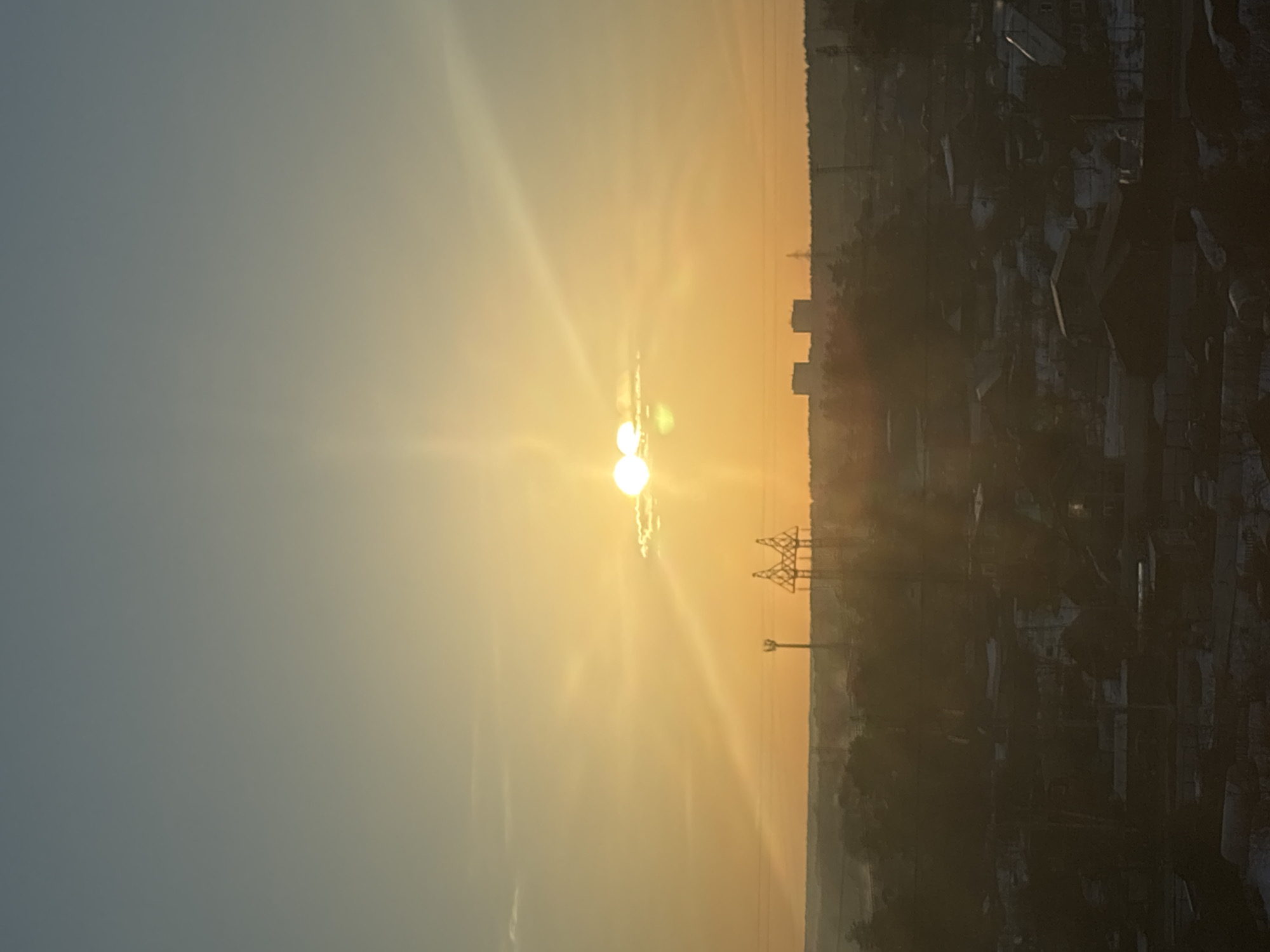 The image captures a sunrise in or near Chaikovsky, Russia. The sun is the central focal point, radiating a bright, yellowish-white light. High-voltage power lines are silhouetted against the bright sky, situated slightly to the right of the sun's position. A skyline of buildings and urban structures is visible in the background, appearing dark and indistinct. The sky is a gradient, transitioning from a light blue on the left to an orange hue closer to the sun. The overall environment is urban or peri-urban, with a suggestion of industrial infrastructure. No people are visible in the frame.