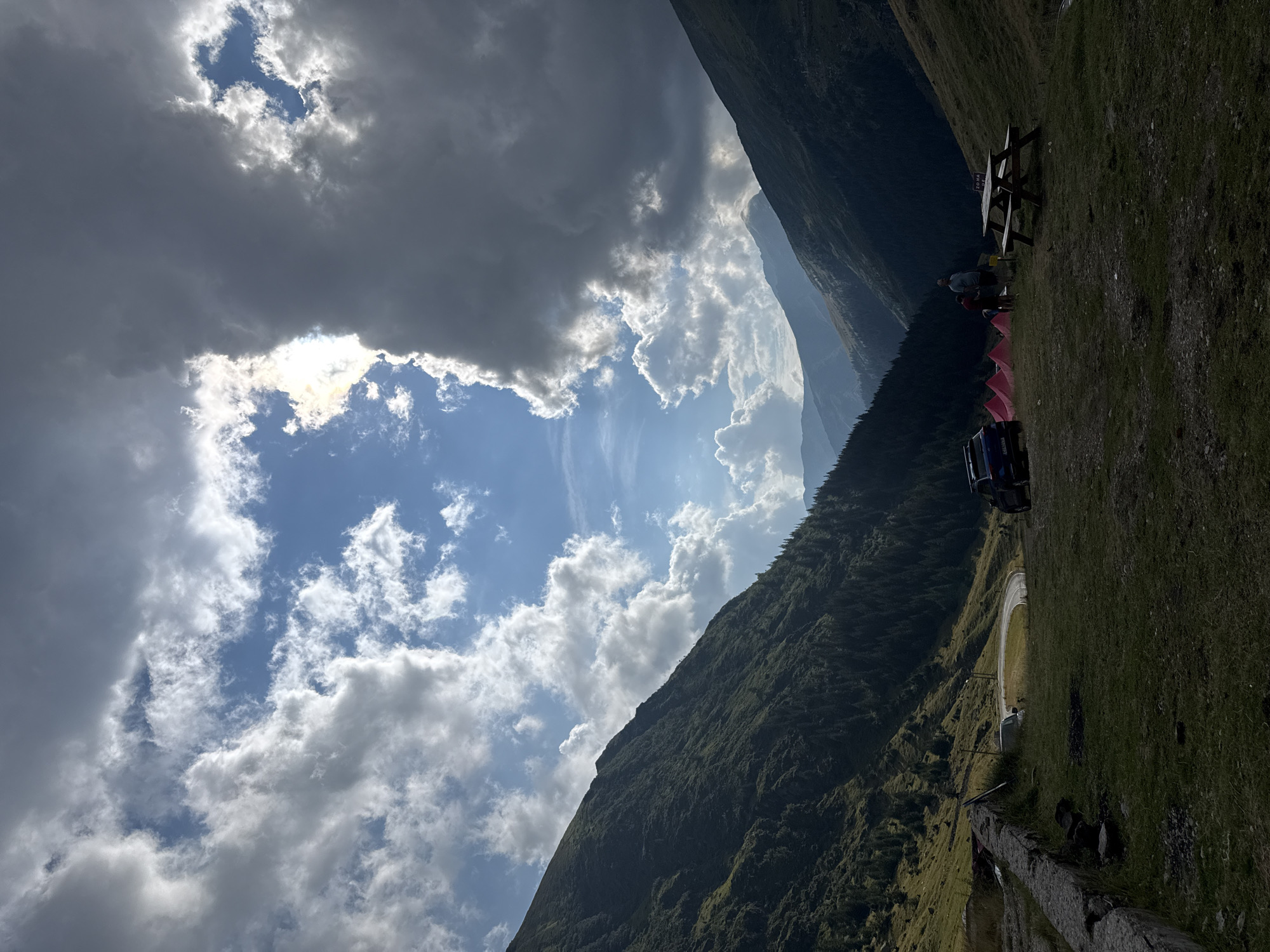 The image depicts a mountainous landscape in Arefu, Romania, with a campsite set up on a grassy slope. The sky is dramatic, with a mix of dark, heavy clouds and patches of bright blue sky with sunlight breaking through. The foreground shows a picnic table, several pink tents, a dark blue SUV, and two individuals standing near the tents. Further back, a winding road is visible, bordered by steep, tree-covered mountains. A stone wall runs along the left side of the foreground. The overall impression is of a remote, rugged natural environment with evidence of human presence.