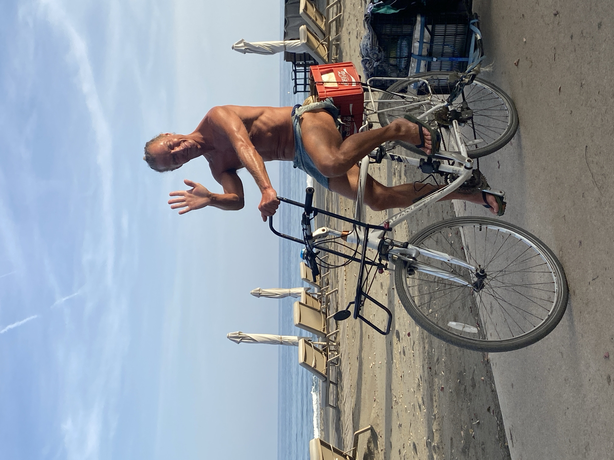 A man is posing on a bicycle on a beach. The man is shirtless and wearing denim shorts. He is smiling and waving at the camera, with his right arm extended. The bicycle is white and has a red Coca-Cola crate attached to the back. The scene is a beach with lounge chairs and umbrellas. The ocean is visible in the background. The sky is blue with some scattered clouds. The overall mood is relaxed and cheerful. The image appears to have been taken during the daytime, likely on a sunny day. The man's tanned skin suggests he is in a warm climate. The presence of lounge chairs and umbrellas indicates a tourist or recreational area. The Coca-Cola crate might suggest a vendor or someone preparing for a refreshment break. The image was taken in Estepona, Spain.