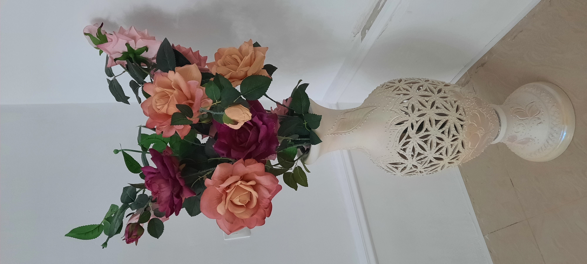 A vase filled with artificial roses stands on a tiled floor, leaning against a white wall. The vase is a light, creamy color and has an ornate, cut-out pattern around its body, resembling latticework. There are also embossed floral designs on the base of the vase. The roses are a mix of colors, including pink, peach, and deep pink, with green leaves. The scene appears to be indoors, likely in a home or commercial setting. The lighting suggests it could be daytime, but there are no specific weather or time-of-day cues. The location is identified as Gudermes, Russia. No people are visible in the image, and there is no visible text.