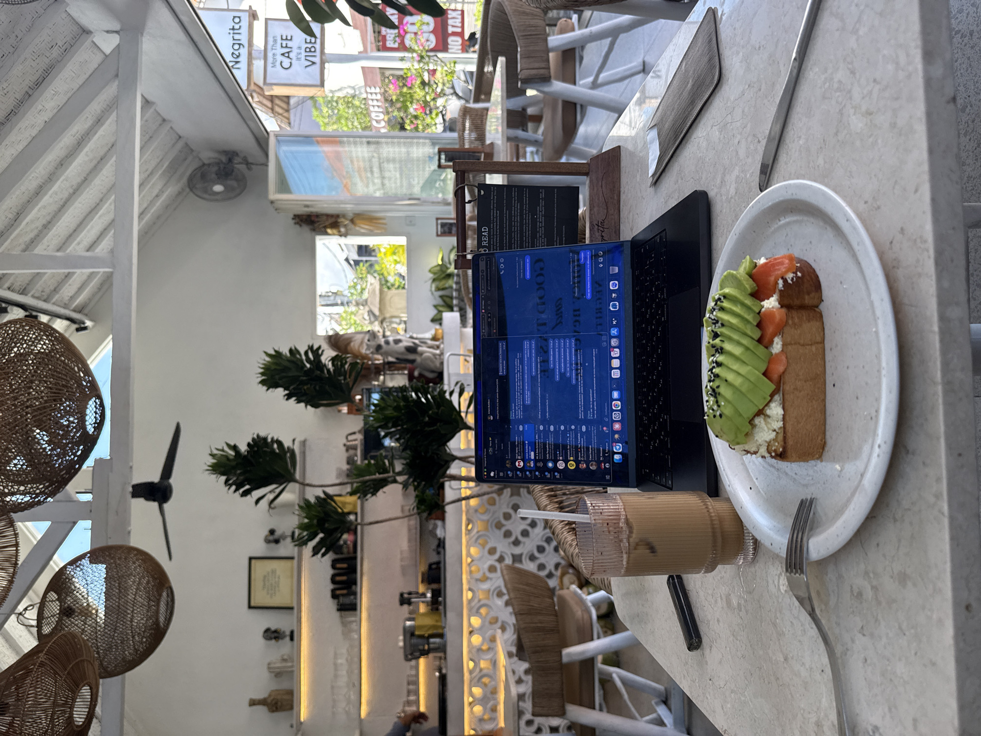 The image presents a bright, daytime scene inside a cafe located in North Kuta, Indonesia. The perspective is an overhead shot of a light-colored table where a black laptop is open, its screen displaying what appears to be a chat or work interface with various icons and text, including the words "FOOD", "GOOD", and "READ". Beside the laptop, a white plate holds a slice of toast topped with layered avocado, cream cheese, and salmon, sprinkled with black sesame seeds. A fork rests on the plate, and a knife is placed next to it. A tall glass containing an iced beverage with a straw is also on the table.
The cafe interior features white wooden beam ceilings and decorative wicker elements, including hanging lights. In the background, a large window reveals a street scene with buildings and greenery. Several signs are visible outside, bearing text such as "Negrita", "More Than CAFE It's a VIBE", "COFFEE", and "NO TAX". Inside, a cafe counter area is partially visible with plants and other decor, along with a small wall-mounted fan. The scene depicts a relaxed setting, suggesting an individual working or enjoying a meal and drink in the comfortable cafe environment.