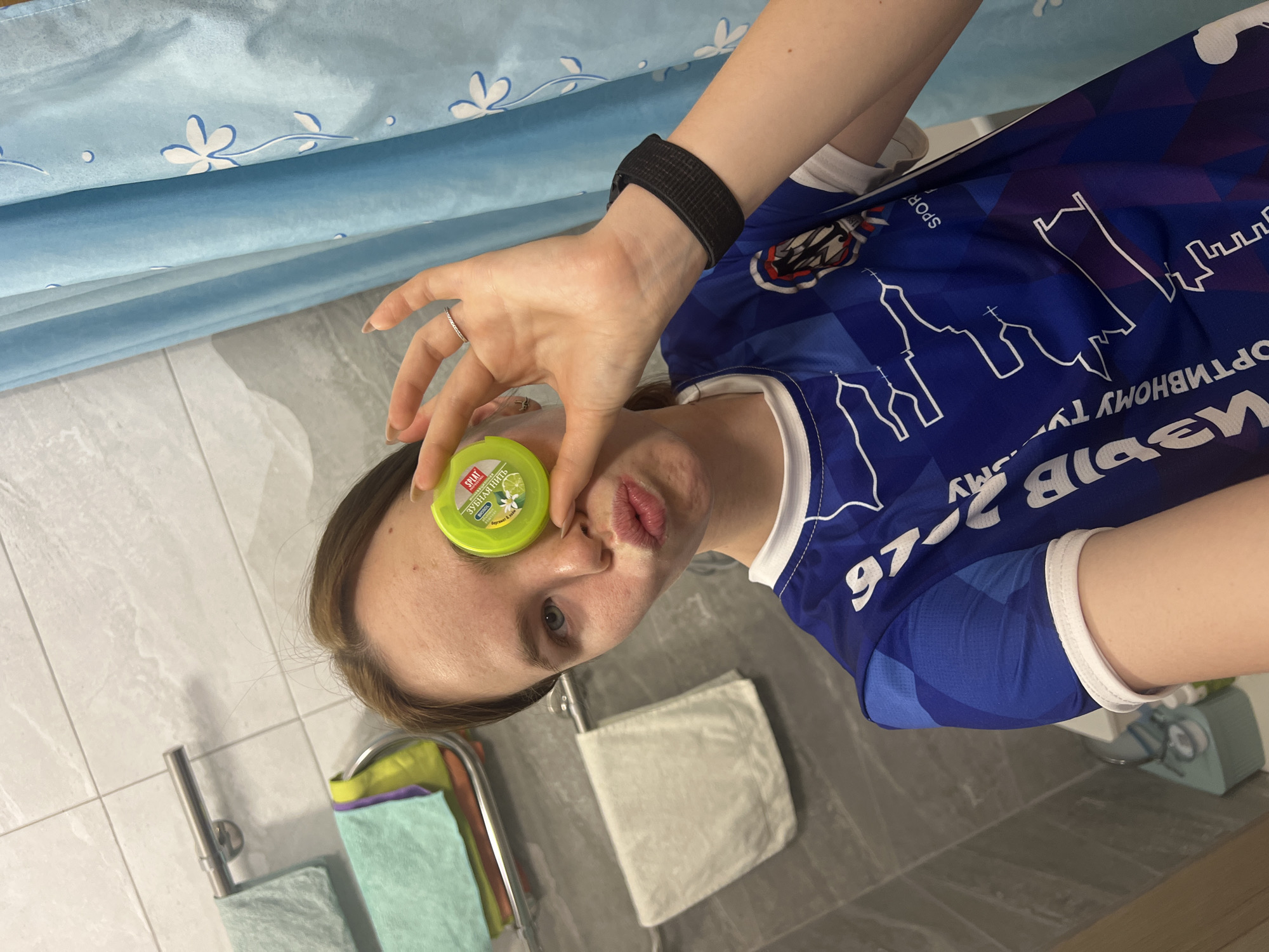 A young woman is holding a container of Splat brand dental floss in front of her eye. She is wearing a blue soccer jersey with white trim. The jersey features a cityscape design and the text "PERMY 2026". This likely indicates that the photo was taken in Perm, Russia. She is also wearing a black activity tracker on her wrist and a ring on her finger. The background includes a blue shower curtain, a tiled bathroom floor and wall, and towels hanging on a rack.