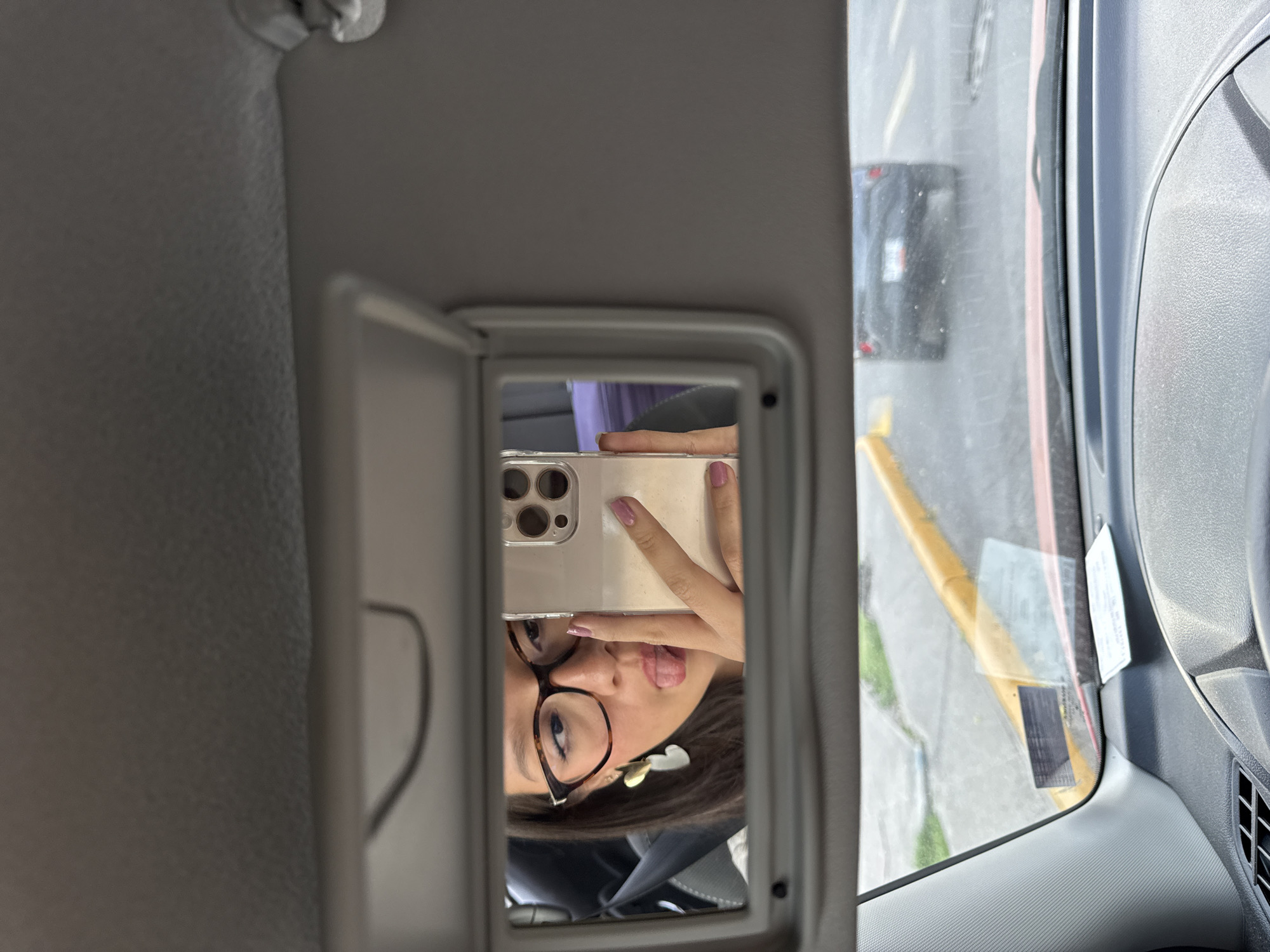 A person is taking a selfie in a car mirror. The person is wearing glasses and has a flower in their hair. The phone they are using has three camera lenses. Outside the car window, part of a street in San Bernardino Tlaxcalancingo, Mexico is visible. The street has a yellow curb and another vehicle.