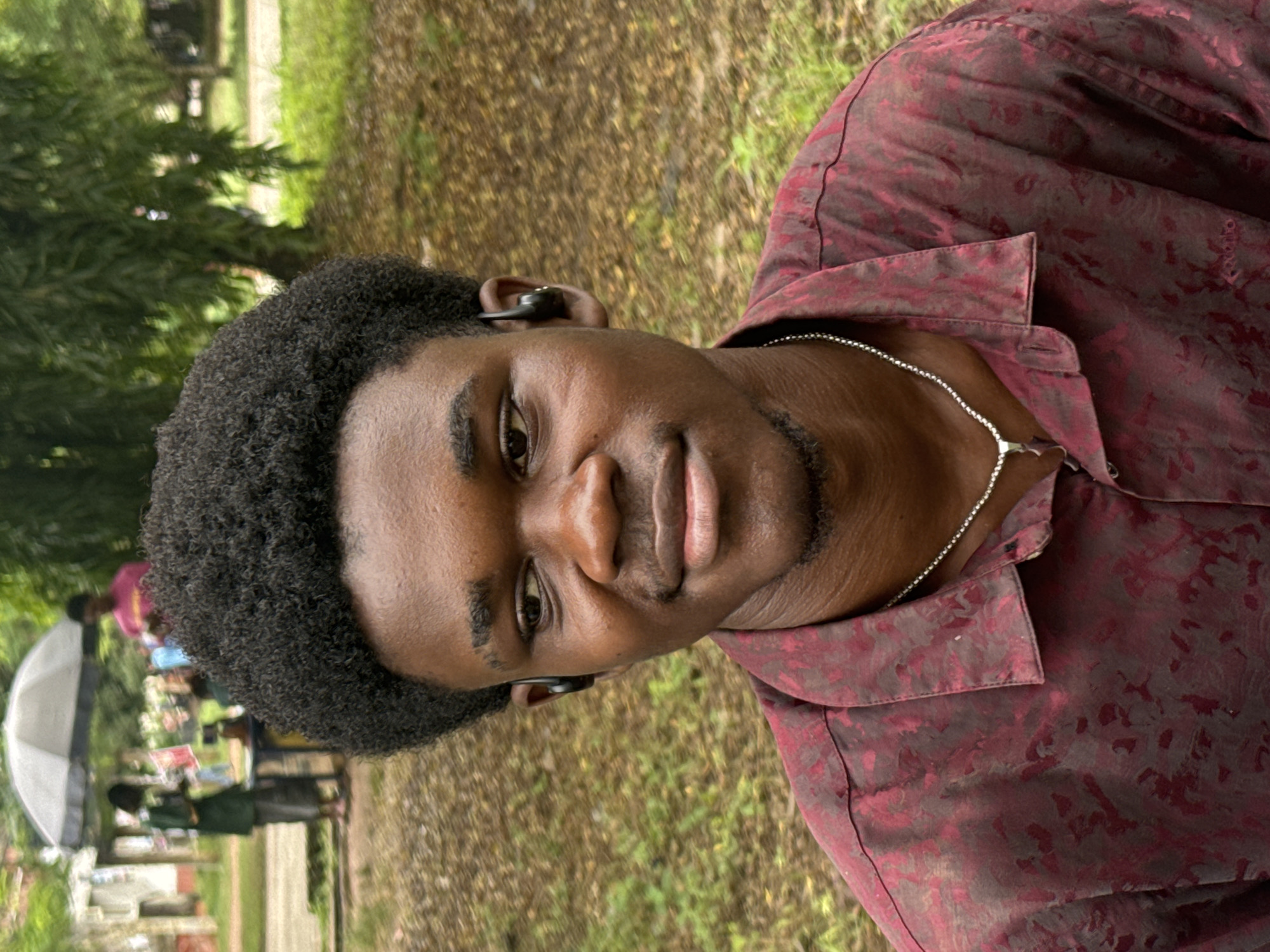 A young Black male with dark, curly hair and a goatee is seated in what appears to be an outdoor setting, possibly in Ihiagwa, Nigeria, given the context provided by the prompt. He is wearing a red, patterned short-sleeved shirt and a silver chain necklace. He has a subtle smile and is looking slightly to his left. There is a black earbud in his left ear. The background consists of lush green grass, scattered leaves, and trees. In the far background, to the left, there are other individuals and a white umbrella, suggesting a public area like a market or a gathering. The lighting suggests it is daytime, possibly overcast or with dappled sunlight.