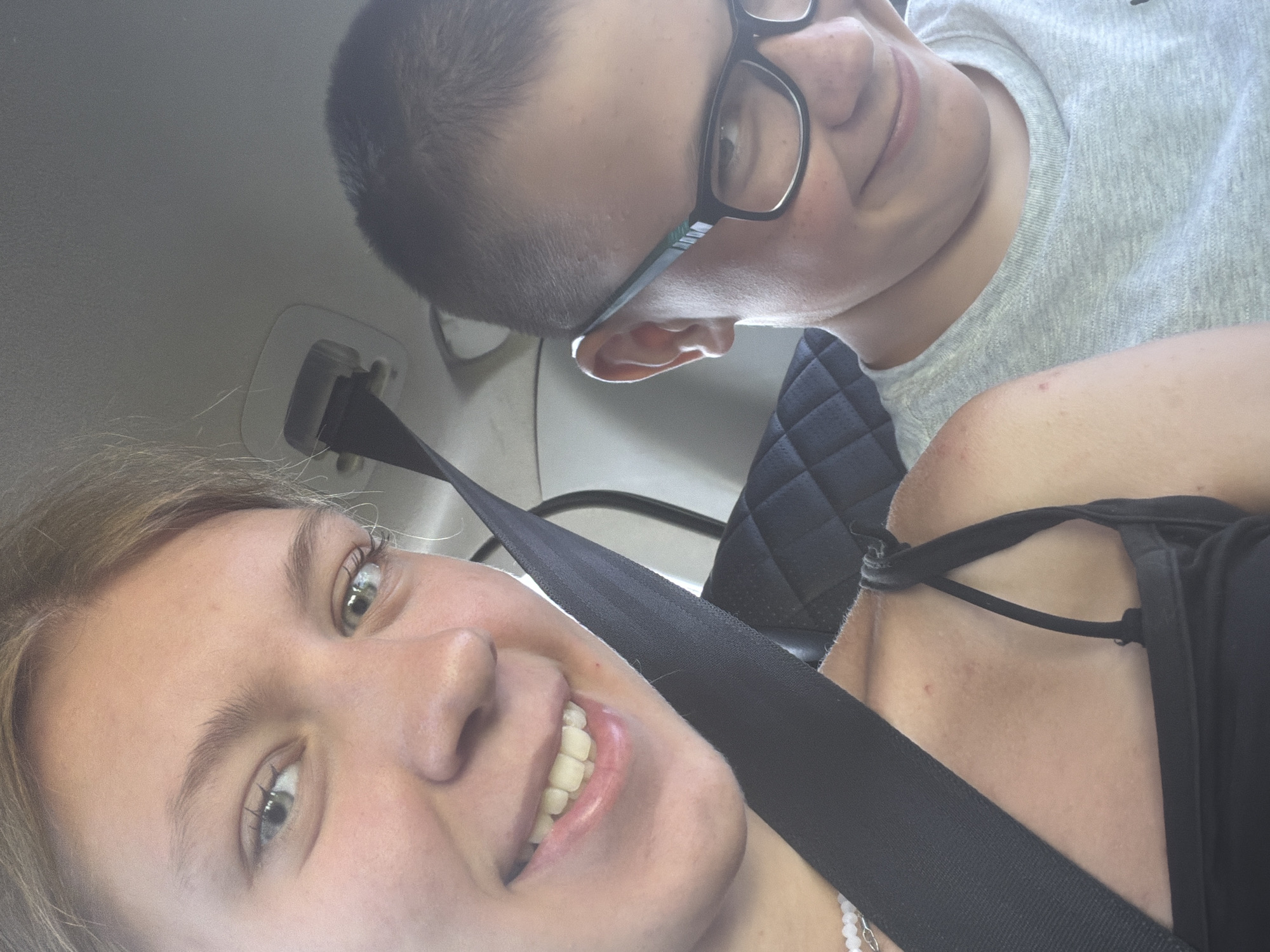The image captures two individuals in the backseat of a vehicle, likely in Novoe Feizullovo, Russia. The person in the foreground, on the left, is a young female with blonde hair and light-colored eyes, smiling directly at the camera. She is wearing a black tank top and a beaded necklace. A black seatbelt is prominently visible, diagonally across her chest. To the right, a young male wearing glasses and a grey t-shirt is partially visible, also smiling. He has short dark hair. The interior of the car is dimly lit, with the textured pattern of the car seat visible behind the subjects. The overall impression is a candid snapshot taken during a car journey.