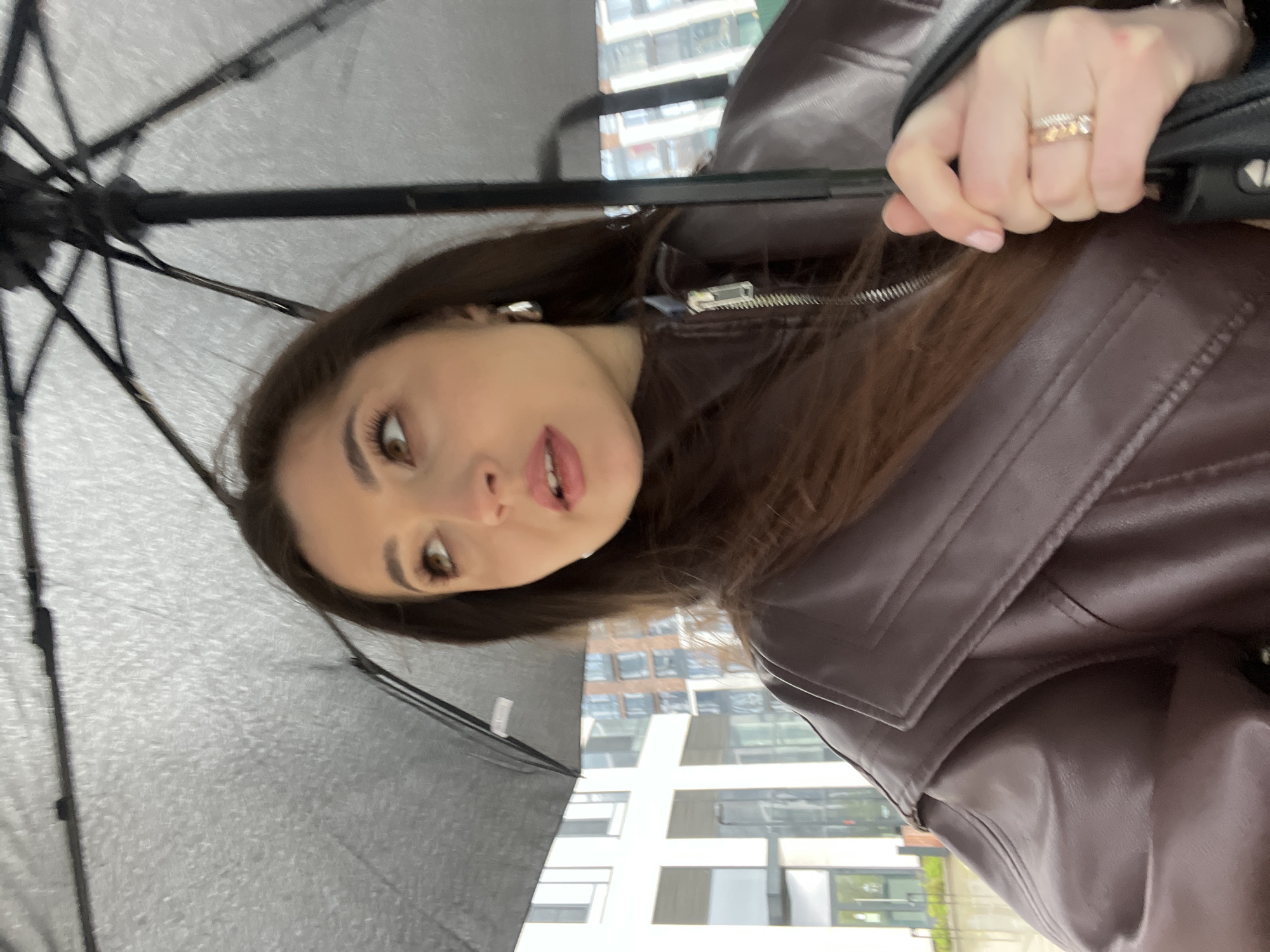 A woman holds a black umbrella outdoors in an urban environment. She wears a dark brown leather jacket. A multistory building is visible in the background.