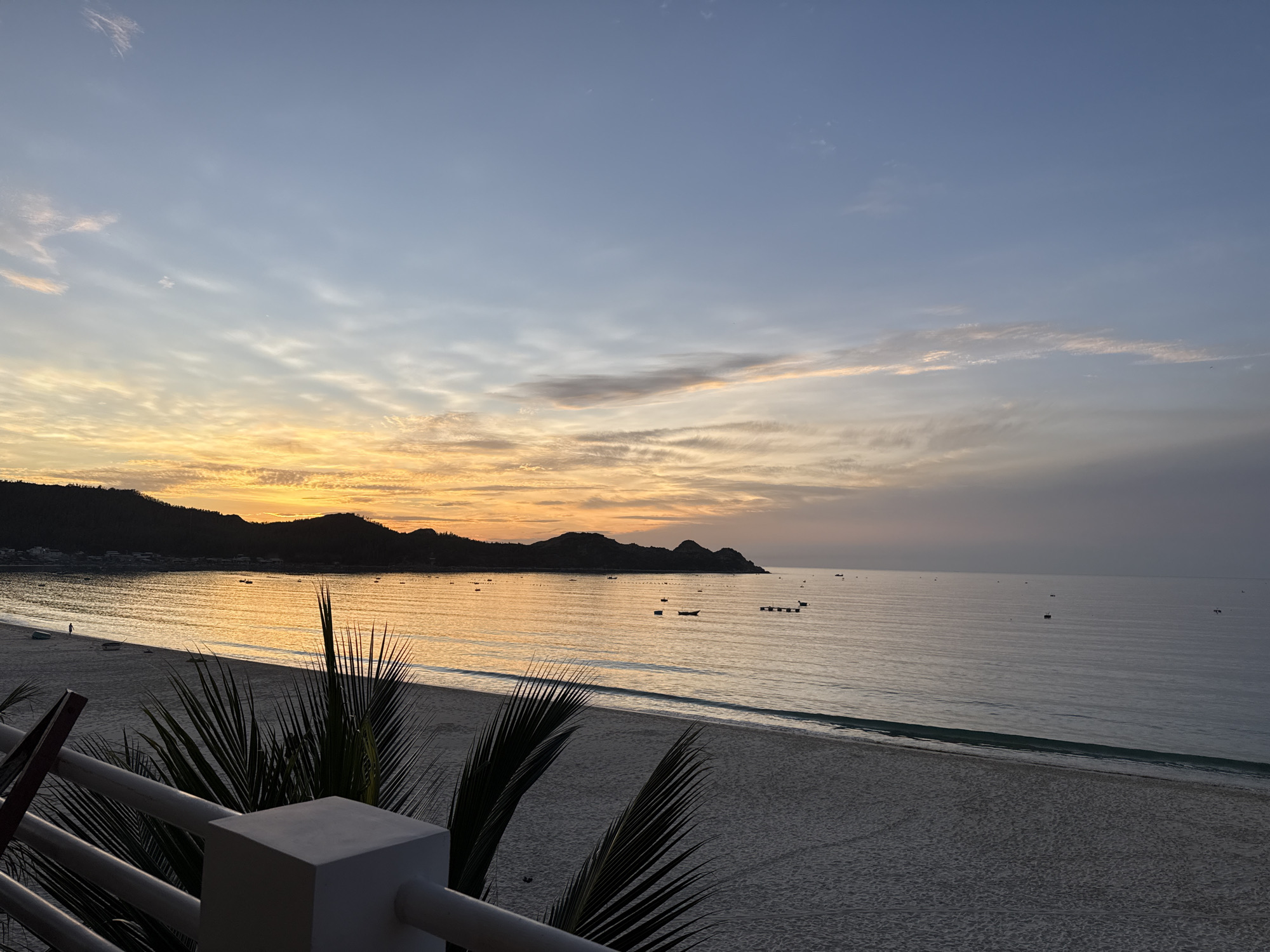 The image captures a serene sunset over a beach in Lệ Uyên, Vietnam. The sky is a canvas of warm oranges and soft blues, with wispy clouds streaked across the horizon. The sun, low in the sky, casts a golden glow on the tranquil sea and the sandy shore. Several small boats are scattered across the water, adding a touch of life to the otherwise still scene. In the foreground, the silhouette of palm fronds frames the view, hinting at the tropical location. The beach itself appears mostly deserted, with a single figure walking near the water's edge. The overall atmosphere is one of peacefulness and natural beauty.