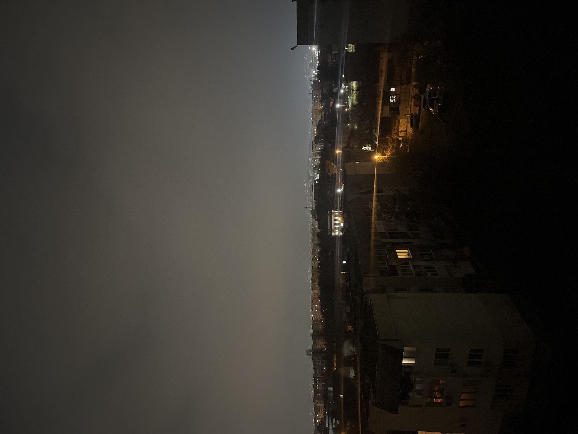 The image depicts a nighttime cityscape in Sofia, Bulgaria, viewed from an elevated position. The left side of the image shows a dark, overcast sky. The right side is dominated by buildings, some with illuminated windows. Streetlights line the roads and illuminate the urban area. Several cars are parked along the street.