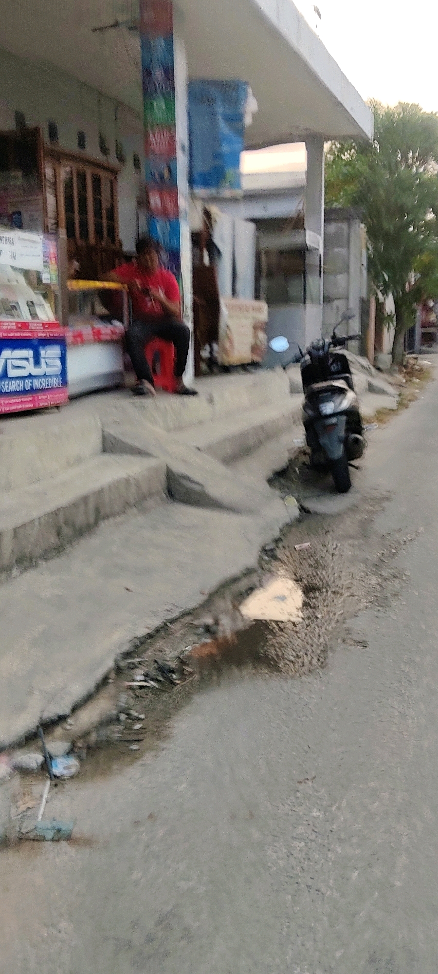 The image shows a street scene in Mekarmukti, Indonesia. In the foreground, a dark-colored scooter is parked on the right side of the street. The scooter is facing towards the viewer. To the left of the scooter, there are concrete steps leading up to a building. On the steps, a person wearing a red shirt and black pants is sitting on a red stool. The person is looking to the left. In the background, the building appears to be a shop with a sign that says "ASUS SEARCH OF INCREDIBLE" in large blue letters. There are also shelves with products displayed inside the shop. A tree is visible on the right side of the image, behind the building. The ground in front of the shop is littered with debris, including plastic bags and other trash. The overall scene suggests a commercial area with some signs of neglect.