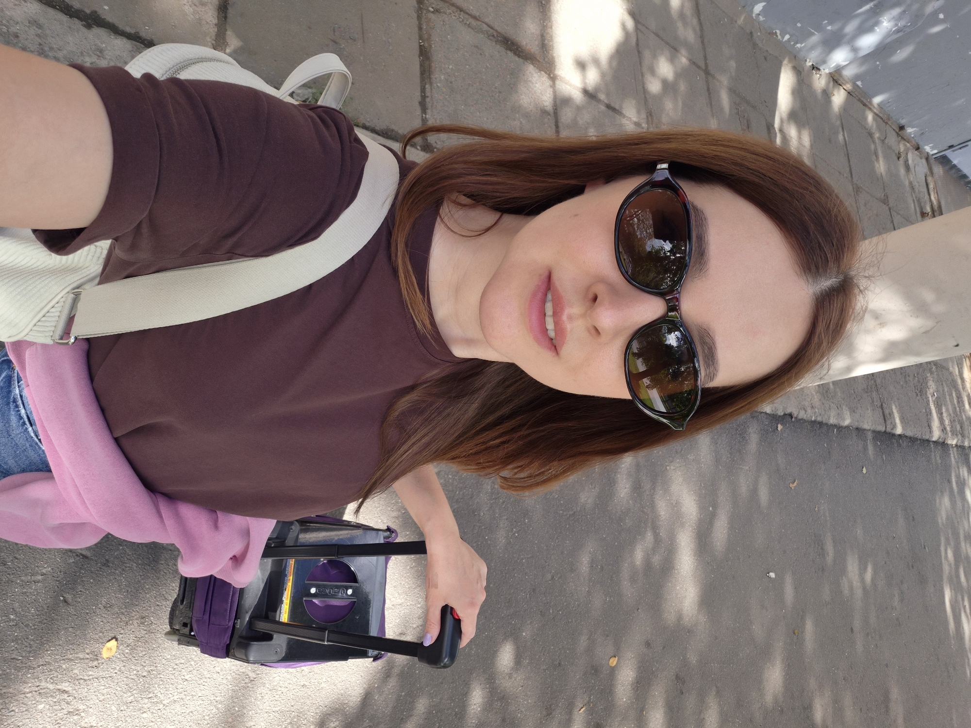 In Saint Petersburg, Russia, on a sunny day, a woman with reddish-brown hair, wearing sunglasses, a brown t-shirt, and blue jeans, is taking a selfie while standing on a paved sidewalk next to a road. She is holding the handle of a small purple suitcase with black accents, and a light-colored bag is slung over her shoulder. A pink garment is draped over her arm. The background shows a grey wall and shadows cast by trees on the pavement and road.