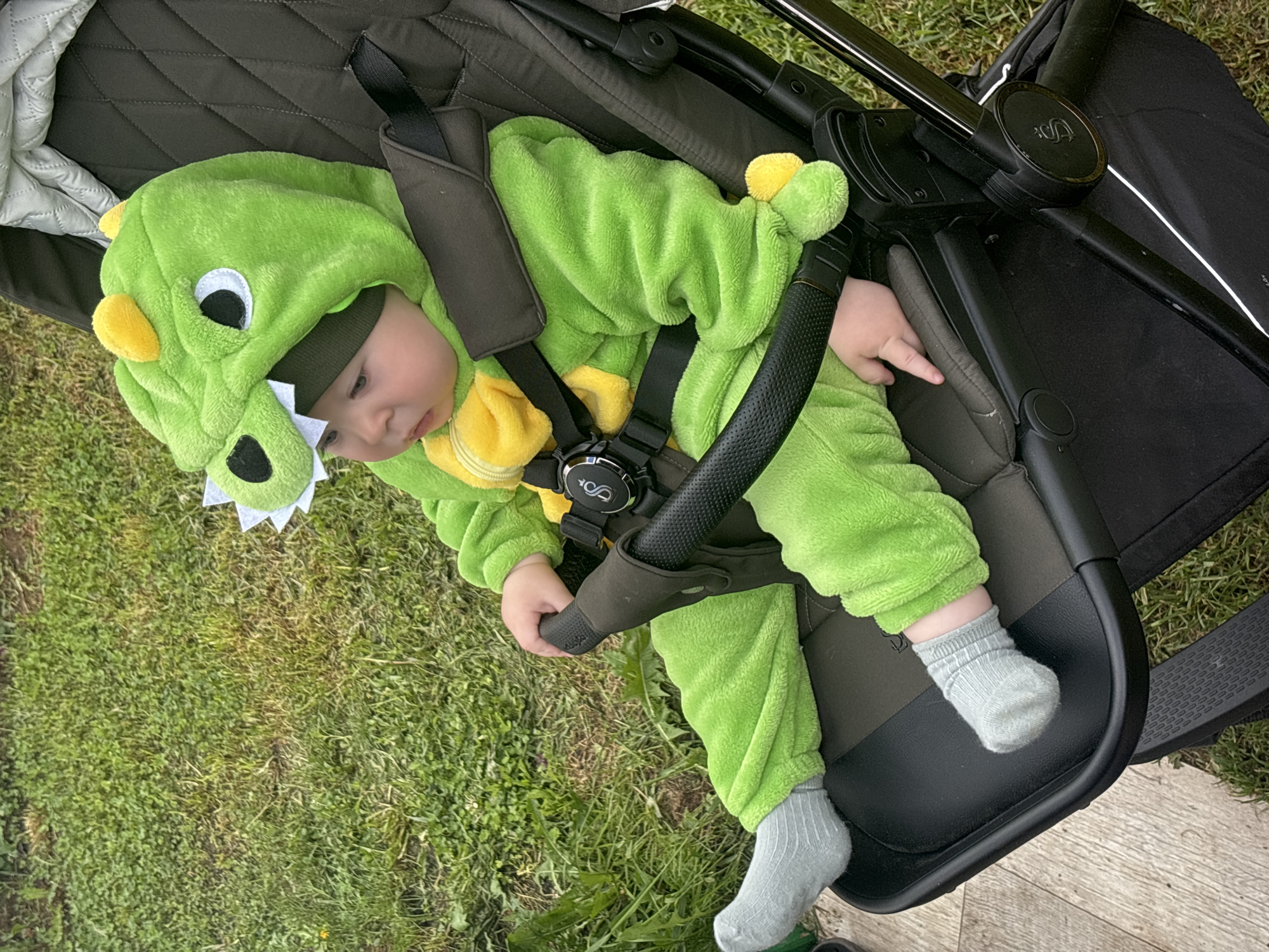The image features a baby dressed in a bright green, plush dinosaur costume, complete with a hood featuring cartoon eyes, soft spikes, and white teeth details. The baby is seated or reclining in a dark-colored stroller, secured by a harness. The buckle of the harness displays the "JOOLZ" logo. The stroller also has a black safety bar and a footrest where the baby's light grey socks are visible.
The scene is set outdoors during daytime, with natural light illuminating the subject. Lush green grass occupies much of the background and foreground, suggesting a garden, park, or yard. In the lower right corner, a section of wooden decking or flooring is visible. The baby appears calm, looking downwards. The image is rotated approximately 90 degrees clockwise from its natural orientation. No specific activity is occurring beyond the baby resting in the stroller.