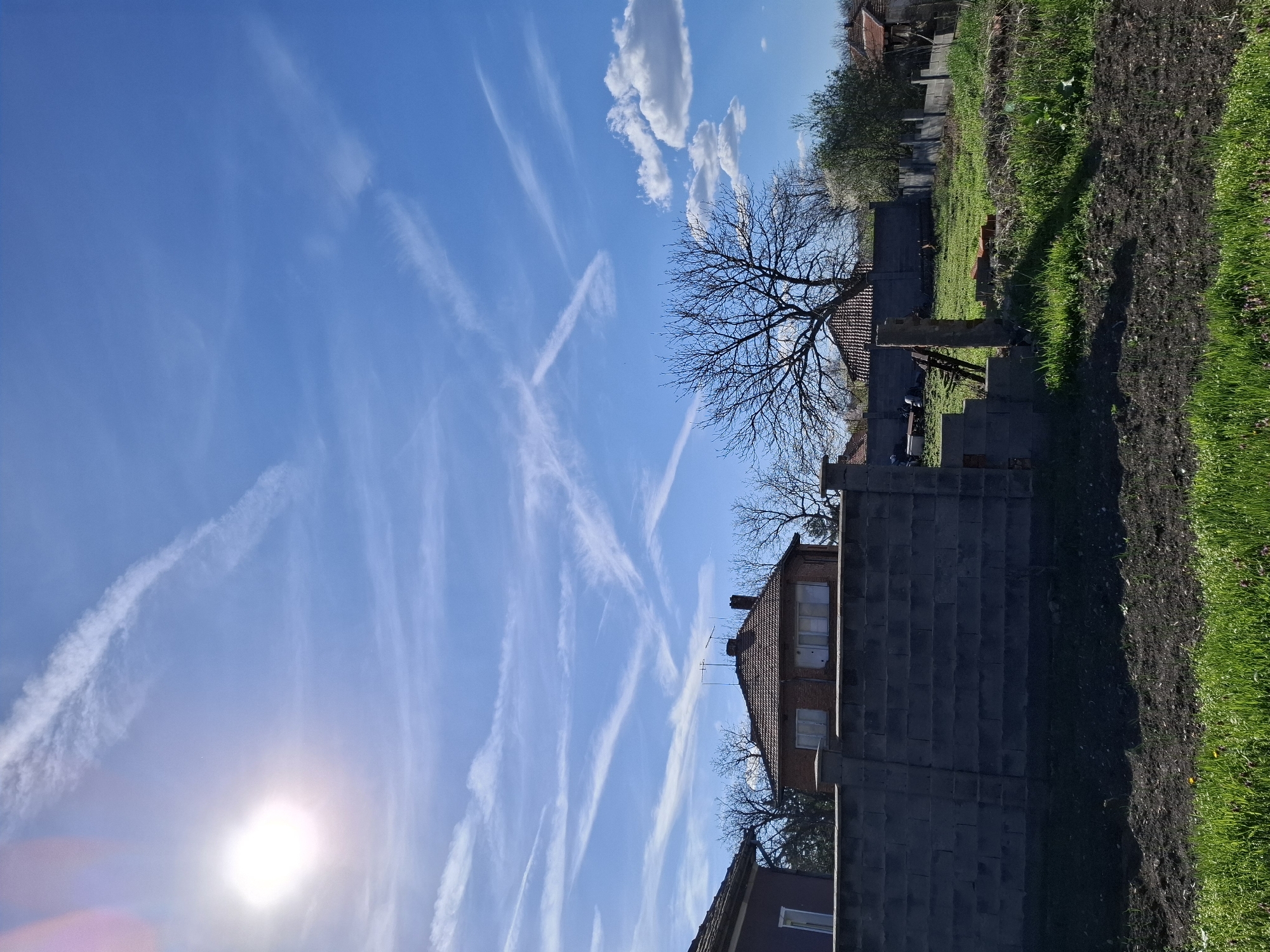 The image depicts a sunny day in Gramada, Bulgaria, with a view of a blue sky filled with contrails. A brick house with a red tile roof and an antenna is visible. To the right of the house is a grey brick wall. A bare tree is positioned near the house. Green grass and tilled soil occupy the foreground on the right side of the image. Other houses and greenery are in the background.