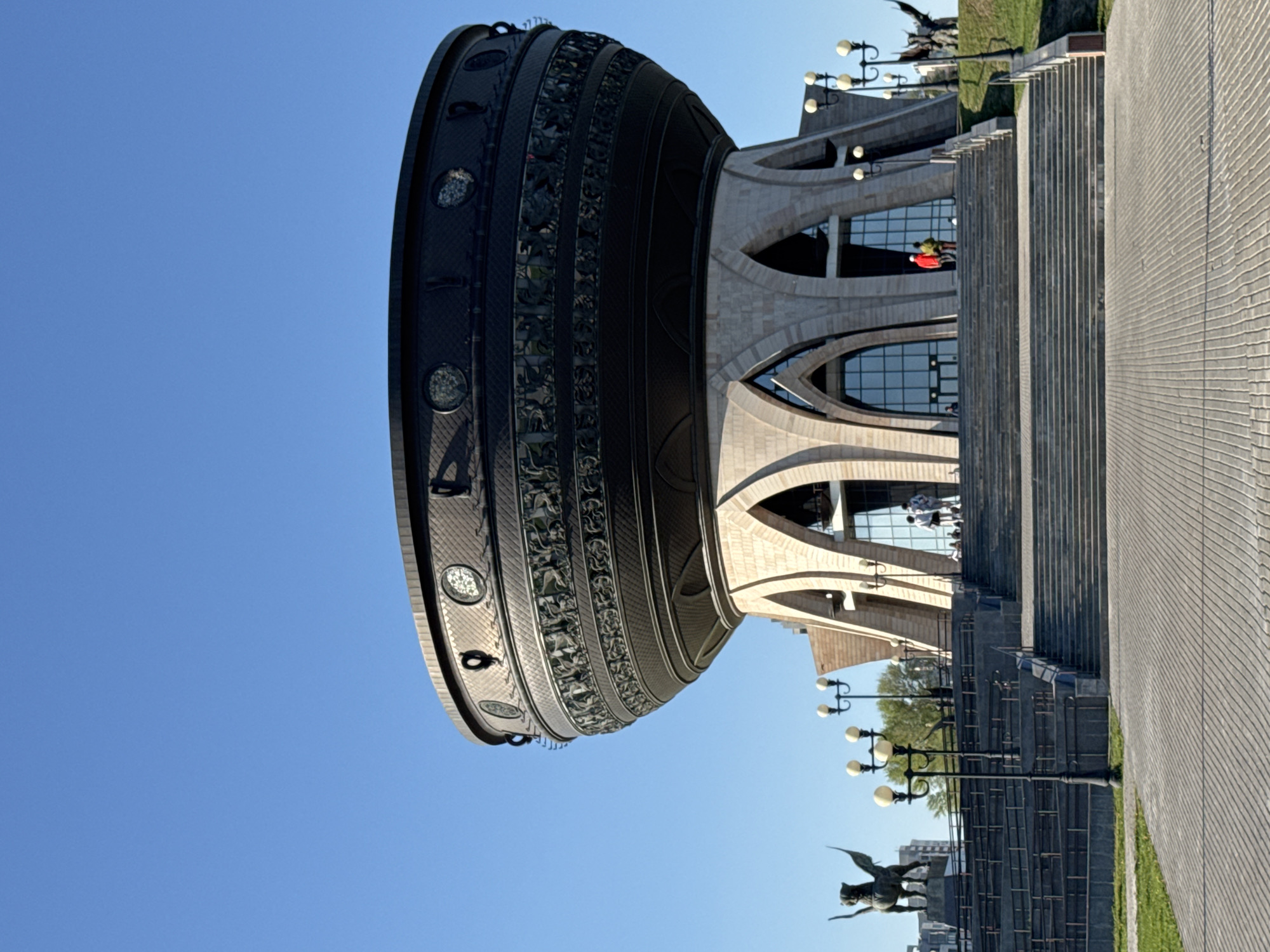 The image shows the Kazan Family Center in Kazan, Russia, against a clear blue sky. The building features a large, cauldron-shaped structure with decorative elements, connected to a sandstone-colored building with arched windows. In the foreground are stairs leading up to the building, a paved walkway, and a statue of a winged creature on a pillar near a lamp post. Two people are visible inside the building, wearing red and green clothes, respectively.