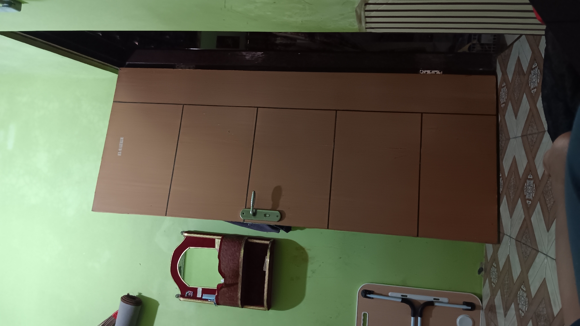 The image depicts an interior scene with a light green wall and a brown wooden door. On the left side of the image, a rolled-up grey mat leans against the wall, and below it is a red and gold shelf with a mirror and small compartments. A silver door handle is attached to the brown door, which is ajar, revealing a darker interior. The floor is tiled with a geometric pattern of beige, white, and brown squares. In the foreground, a person's legs and arm are visible, suggesting they are lying down or sitting on the floor. A portion of a beige laptop table with black legs is also visible on the left. The scene appears to be a room in a residential setting, likely in Sonobekel, Indonesia, given the context of the prompt.