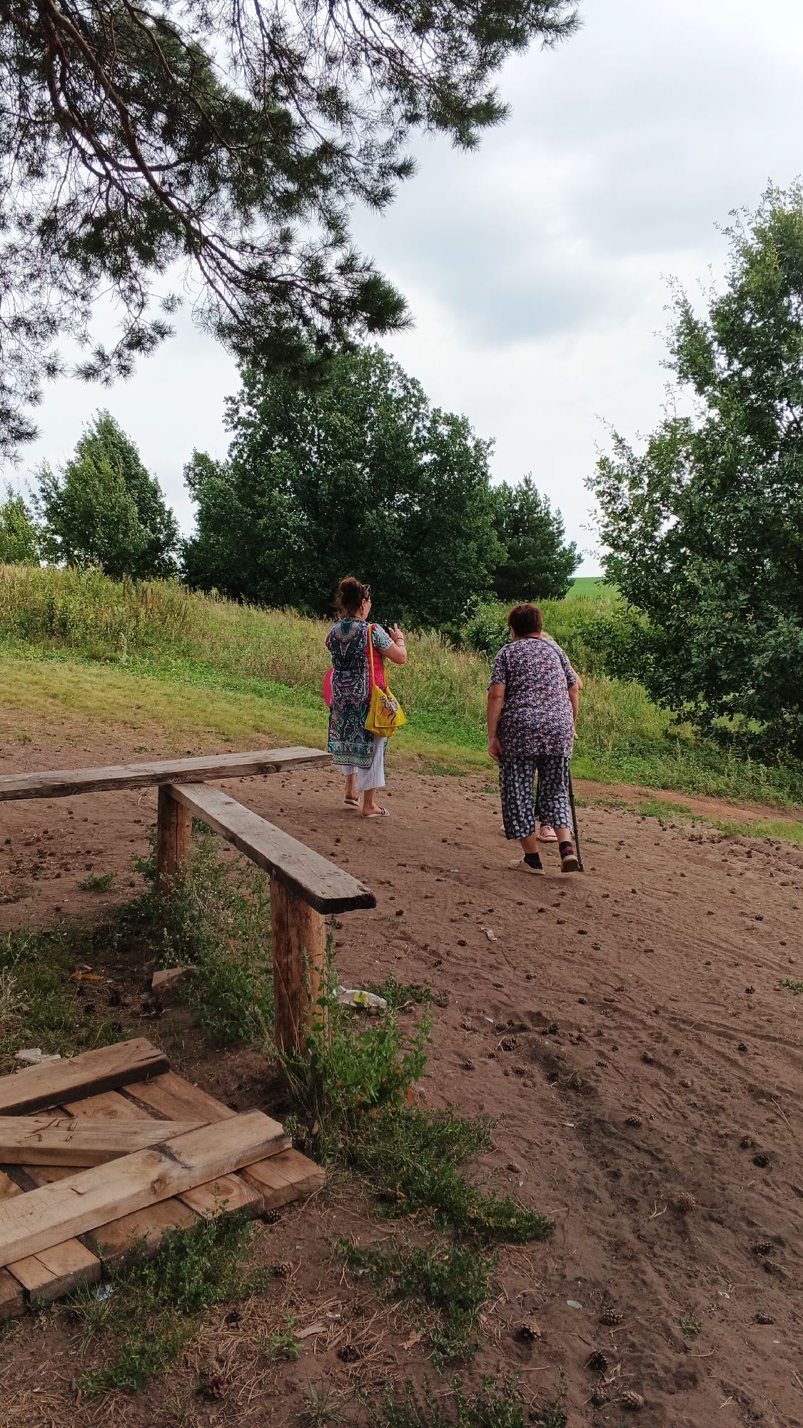 Two women are walking on a dirt path in a rural area. The woman on the left is wearing a patterned dress and carrying a yellow bag. The woman on the right is wearing a floral patterned outfit and using a cane. A wooden bench is to the left of the path. In the background, there are trees and a cloudy sky. The scene is likely in the city of Подгорное, Russia, given the context.