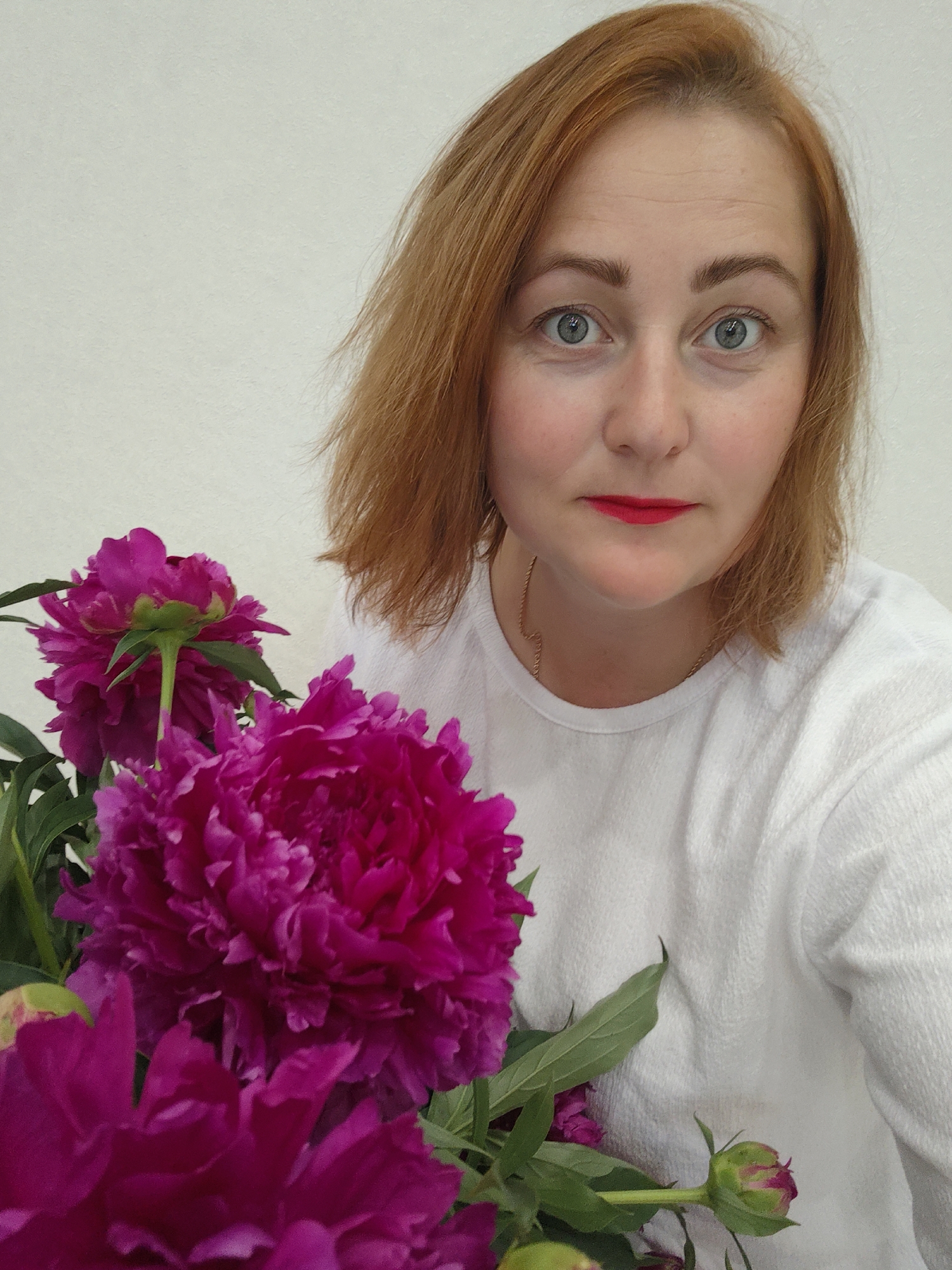 In the foreground of the image, a bouquet of vibrant pink peonies is prominently displayed, with their lush petals filling a significant portion of the frame. Behind the flowers, a woman with auburn hair and light blue eyes is visible, looking directly at the camera. She is wearing a simple white t-shirt and has red lipstick on. The background is a plain, light-colored wall, suggesting an indoor setting. There is no indication of the specific location within Markovskii, Russia.