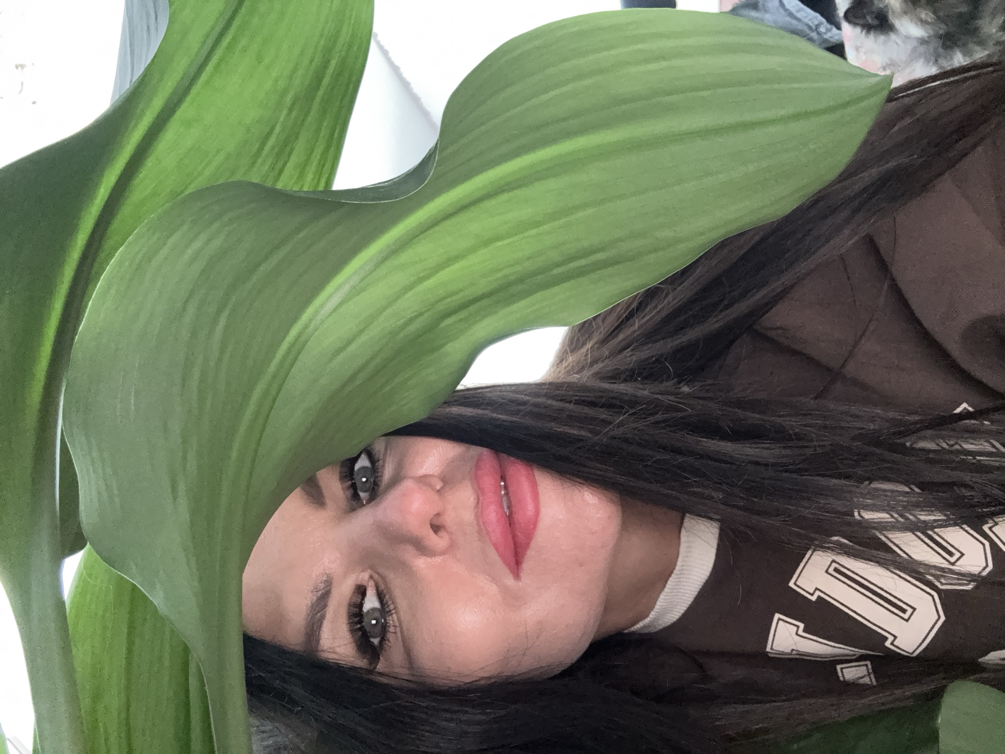 A woman with long dark hair is partially obscured by large green plant leaves. She is wearing a brown shirt with white lettering around the collar, only partially visible. Her eyes are open, and she is looking at the camera. The photograph is rotated sideways. It is not possible to ascertain the location in Dubovaya, Russia from this image.