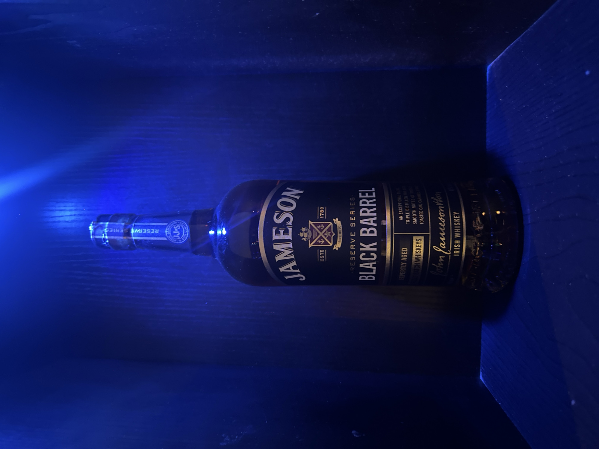 A bottle of Jameson Black Barrel Irish Whiskey stands upright on a dark, wooden surface under blue lighting. The bottle is the central subject of the image, with its label prominently displayed. The label features the Jameson logo, "RESERVE SERIES," "BLACK BARREL," and "IRISH WHISKEY." The background is dark and textured, suggesting a shelf or alcove. The blue lighting casts a strong glow on the bottle and the surface it rests on, creating a dramatic and somewhat mysterious atmosphere. The overall scene is static and focused solely on the product. There are no people or other objects visible. The location in the city of Howayi, Nigeria, is not discernible from the visual information provided in the image.