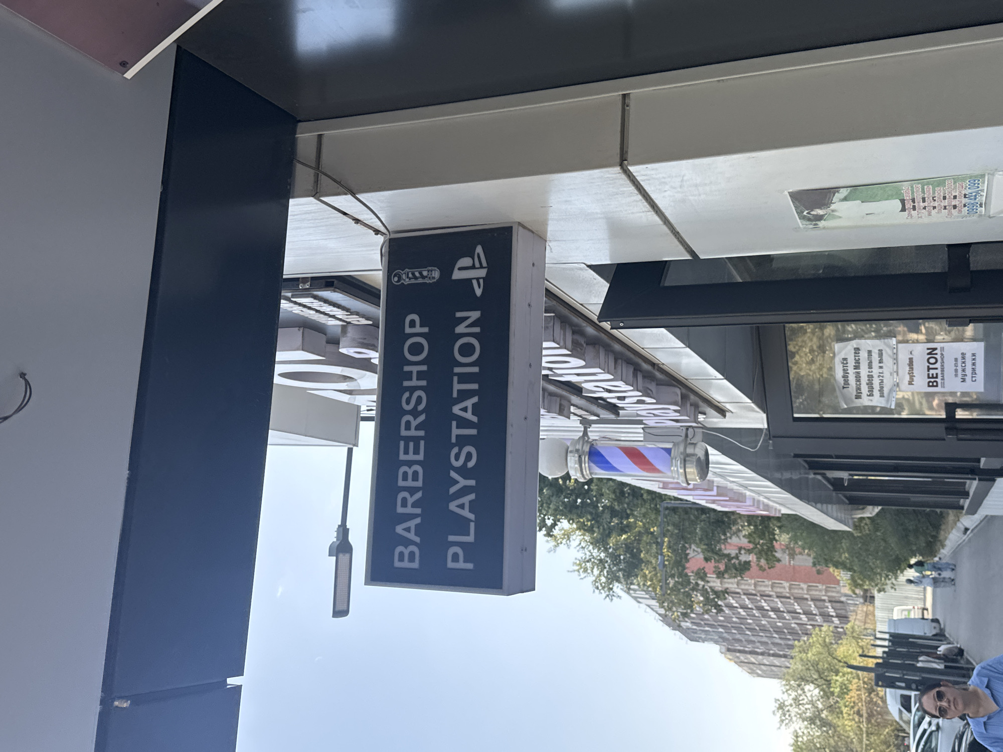 The image shows the facade of a business establishment with a prominent sign reading "BARBERSHOP PLAYSTATION" with a PlayStation logo. Below this sign, a barber pole with red, white, and blue stripes is visible. The building appears to be located on a street with trees and a tall, unfinished apartment building in the background. There are cars parked along the side of the road. A woman wearing sunglasses and a blue shirt is walking towards the camera in the foreground. Signs with Cyrillic text are also present on the building, suggesting the location is in a Russian-speaking region. One sign on the glass door mentions "BETON BARBERSHOP" and "МУЖСКИЕ СТРИЖКИ," meaning "Men's Haircuts." Another sign provides a phone number, "0998 443 099." Based on the presence of Cyrillic script and common business names, this establishment is likely located in Bishkek City, Kyrgyzstan.