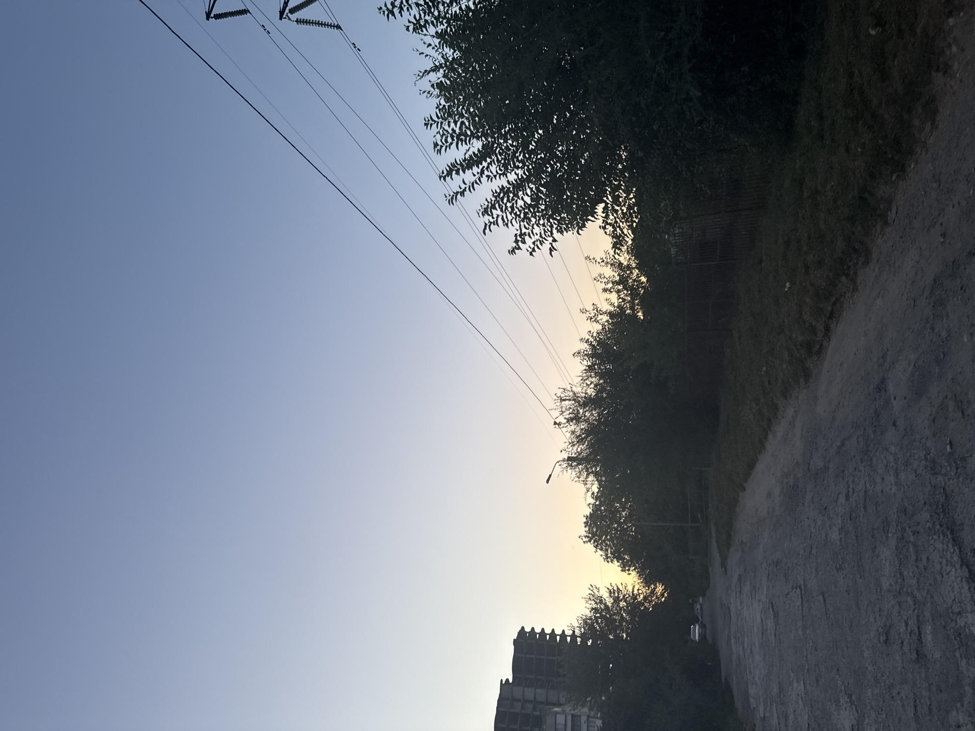 The image depicts a rural or semi-urban road in Zaporizhzhia, Ukraine, during twilight. The sky is a clear gradient of pale blue to light yellow near the horizon, indicating either sunrise or sunset. Power lines stretch diagonally across the upper right portion of the frame, supported by a pylon visible at the very edge. On the left side of the road, a multi-story building under construction, with exposed concrete and scaffolding, stands silhouetted against the sky. The road itself is unpaved and appears to be made of gravel and dirt, with some patches of rougher terrain. Dense foliage and trees line the right side of the road, with a wooden fence partially visible behind them. A lamppost stands among the trees in the mid-ground. In the distance on the left, a white vehicle is parked on the side of the road. The overall atmosphere is serene and quiet, characteristic of the early morning or late evening in a less developed area.