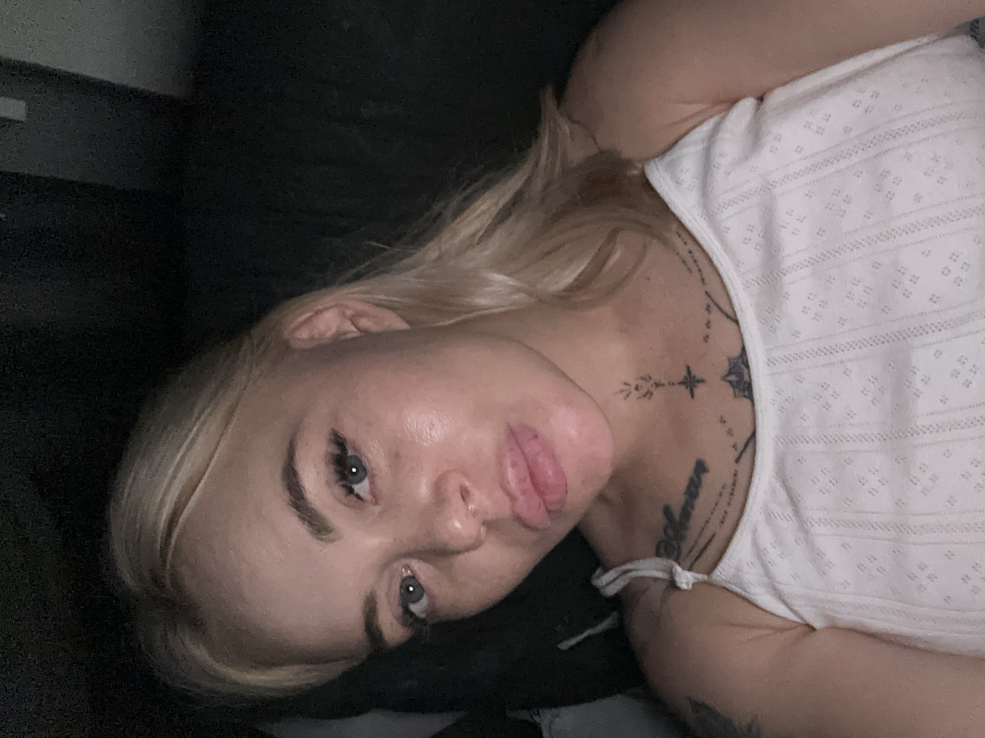 A close-up, high-angle shot depicts a light-skinned woman with blonde hair and dark eyebrows, lying down in a dimly lit indoor setting. Her head is tilted to the left, and her body is partially visible in a white top with a subtle textured pattern. She is looking directly at the camera with her eyes slightly wide open, giving a somewhat intense or curious expression. Her lips appear full and slightly parted.
She has several tattoos on her upper chest and neck area. One tattoo near her right collarbone features a star with radiating lines and possibly some fine script. Another tattoo, partially visible on her left shoulder, also appears to be script or a symbol.
The background is dark and appears to be a solid surface, possibly a wall or furniture, contributing to the intimate and focused nature of the portrait. There are no clear indicators of time of day or specific location, but the low light suggests evening or night. No other objects or people are visible in the frame.