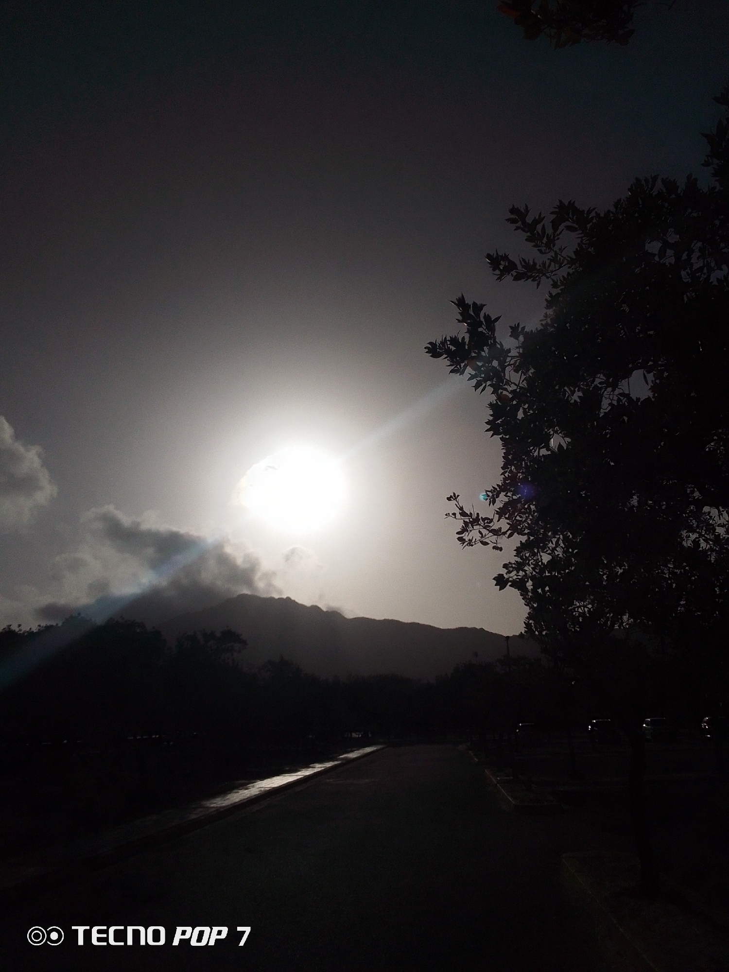 The image captures a landscape at or near dusk in Pedro González, Venezuela. The sky is overcast, with a bright sun partially obscured by clouds and mountain silhouettes in the distance. A ray of sunlight pierces through the clouds, creating a stark contrast between light and shadow. In the foreground, a paved path or road stretches into the distance, reflecting some light. A tree is silhouetted on the right side of the frame, obscuring part of the scene. To the bottom left the camera brand and model name are printed "TECNO POP 7". The scene is predominantly dark, with limited details visible due to the low light conditions.