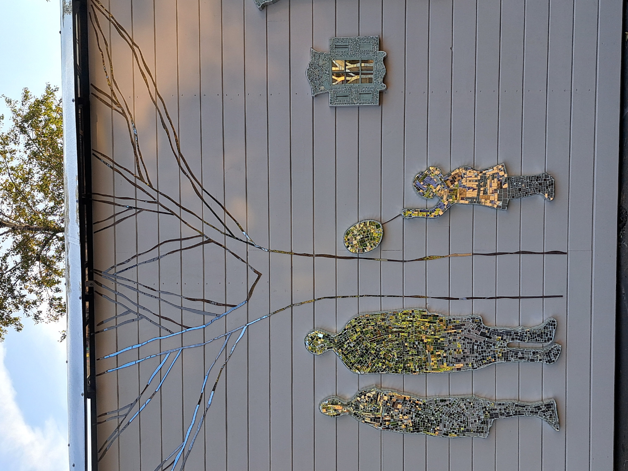 The image depicts an outdoor mural on a horizontal wood-paneled wall. The mural features a mosaic of a tree with bare branches that extend across the upper portion of the wall, some branches appearing metallic and others a darker tone. Below the tree, three mosaic figures are positioned. On the left, two standing adult-sized figures are depicted side-by-side, rendered in mosaic tiles that reflect light. The figure on the right is more densely tiled with green and yellow hues, while the figure on the left is primarily tiled with silver and gray fragments, appearing somewhat incomplete or stylized. To the right of these figures, a smaller mosaic figure of a child is shown holding a balloon. The child is also made of reflective mosaic tiles in various colors. Further to the right on the wall is a decorative mosaic piece resembling a window frame, also with reflective elements. The backdrop of the mural is the light-colored wood paneling, and above the mural, a sliver of a tree canopy and a blue sky with some clouds are visible. The scene appears to be illuminated by sunlight, casting reflections on the mosaic elements. There is no information in the image that indicates the location of this mural in Irkutsk, Russia.