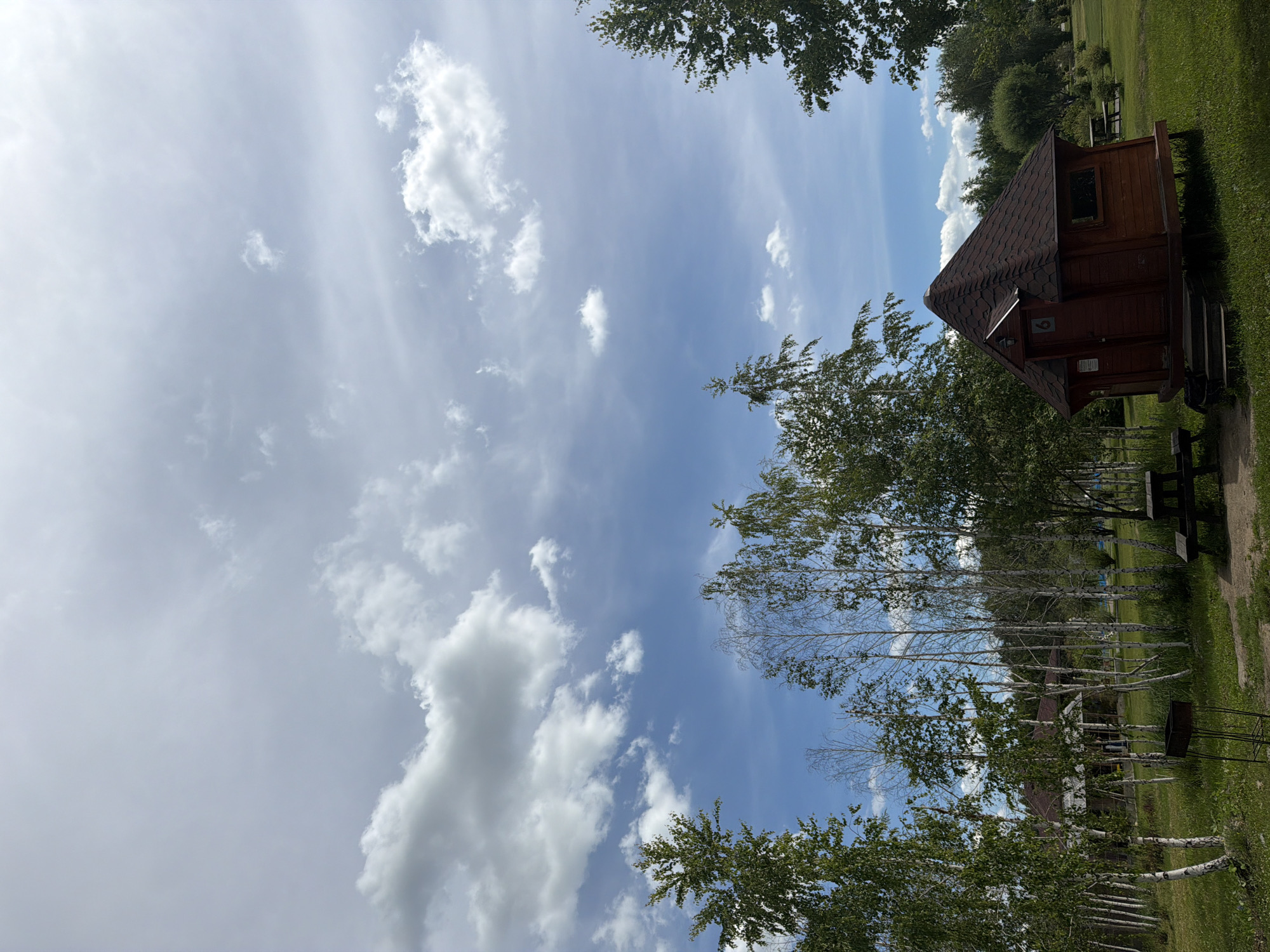 The image captures a scene at a campsite or recreational area, possibly in Kremenkul', Russia, characterized by a small, rustic wooden cabin with a dark red, angular roof. The cabin has a single door with a small number "6" visible and a window on its side. It is elevated on wooden stilts and accessed by a short set of stairs. Surrounding the cabin is a grassy area with several picnic tables, one visible in the foreground and others further in the background.
Several trees, including birch trees with their distinctive white bark and other deciduous trees with green foliage, are scattered throughout the scene. In the distance, a larger building, possibly a house or another facility, is partially visible through the trees. The sky is a mix of blue sky and scattered white clouds, indicating a pleasant, possibly summer day. The overall impression is one of a peaceful, outdoor setting. There are no people or animals visible in the image. The lighting suggests it is daytime, with the sun casting soft shadows.