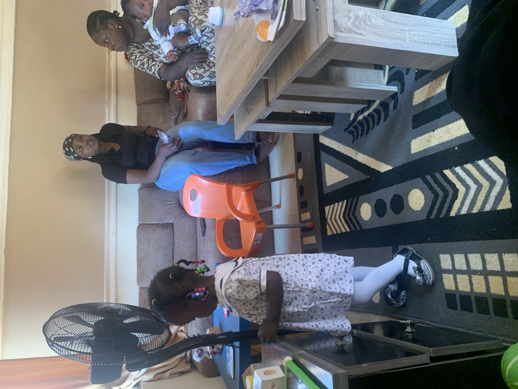 In the city of Gura Top, Nigeria, a scene unfolds within what appears to be a living room or common area. The room is furnished with a brown sofa and a wooden coffee table arrangement. Natural light illuminates the space, casting shadows and highlighting details.
In the foreground, a young girl stands facing away from the camera, her back to a large black oscillating fan. She wears a light-colored dress with a repetitive pattern and white tights, and her feet are clad in black shoes adorned with bows. Her dark hair is styled in braids, accentuated with colorful beads.
Seated on the sofa are two women and a baby. One woman, wearing a black headscarf and a black t-shirt with blue jeans, holds a white object in her hand. Beside her, another woman, dressed in a patterned top, cradles a baby dressed in white. The baby is held securely in her arms.
An orange child-sized chair is positioned between the sofa and a small blue table. The floor is covered by a patterned rug featuring geometric shapes and stripes in shades of black, grey, and yellow. The coffee table is a set of nested tables made of light-colored wood. On the table, various items are visible, including what appears to be a blue item, a white bowl, and some crumpled plastic.
The background consists of a plain, light-colored wall with a white molding strip running across it. A curtain is visible on the far left, partially obscuring the wall. The overall impression is of a casual indoor gathering.