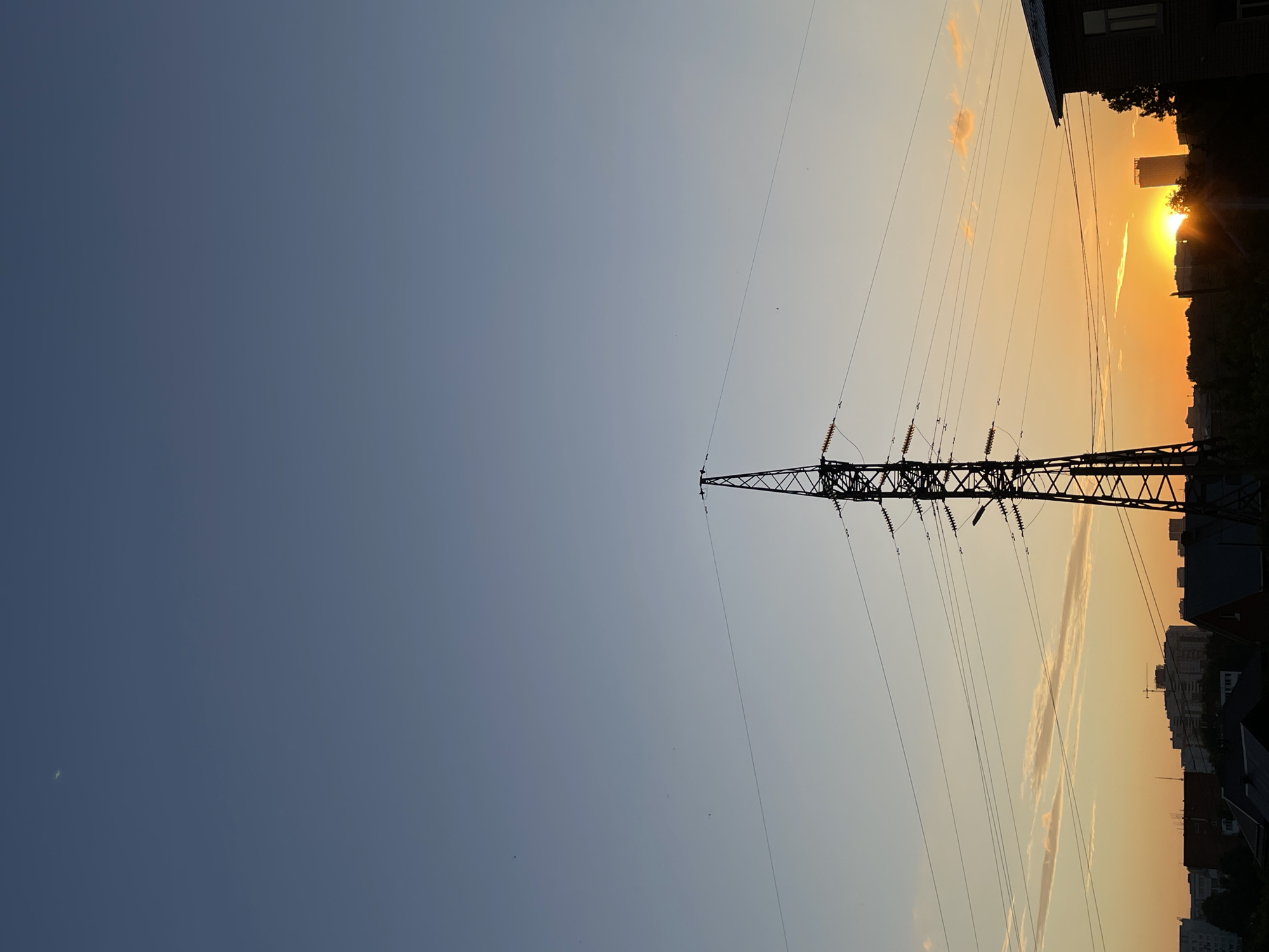The image shows a power line tower silhouetted against a sunset sky in an urban environment. The sky transitions from a deep blue at the top to a warm orange and yellow near the horizon. Wispy clouds are visible. Below the sky, a silhouette of buildings and trees forms the cityscape. The power line tower is the most prominent object in the foreground, with its complex structure of metal supports and wires stretching across the frame. The sun is setting behind a tall building, casting a bright glow. The overall scene captures a moment of transition between day and night in a city.