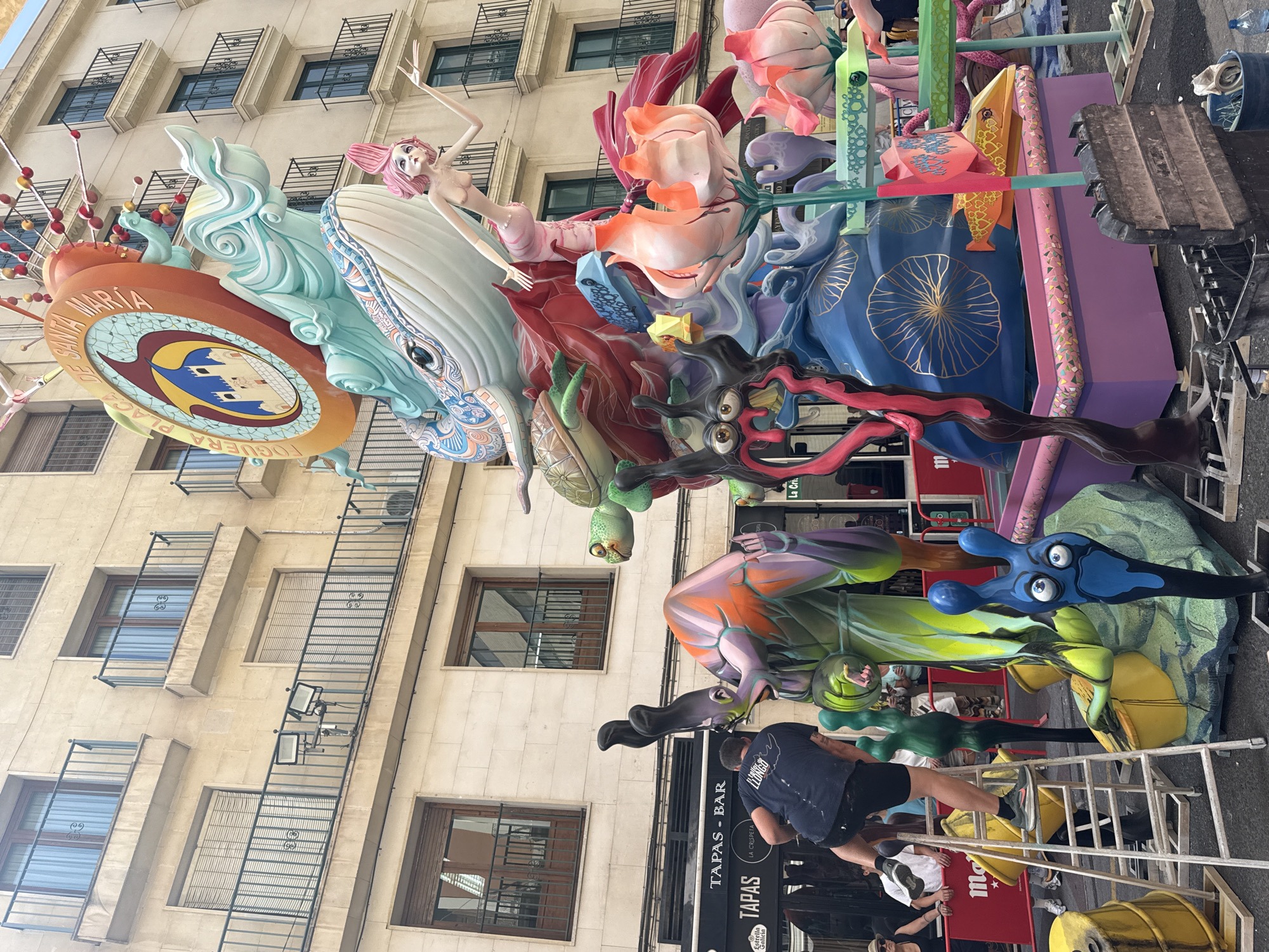 In an outdoor urban setting, a large, colorful, and intricate sculpture is the central focus. The sculpture depicts various fantastical creatures and elements, including a whale, a mermaid, a turtle, and stylized floral motifs, all rendered in vibrant hues of blue, green, orange, pink, and red. A man wearing a dark blue t-shirt and black shorts is standing on a ladder, appearing to be working on or inspecting the sculpture from the side. He is positioned to the left of the sculpture's base. In the background, a multi-story building with a light-colored facade and balconies is visible. Several people are scattered around the scene, some observing the sculpture and others in the background engaged in various activities. Several large yellow barrels are present in the foreground and to the left of the sculpture. Signs for "TAPAS - BAR" and "ESTRELLA GALICIA" are visible on a storefront to the left of the sculpture. The ground is paved, and construction materials, including wooden planks and containers, are present around the base of the sculpture, suggesting it is either under construction or being prepared for an event. The lighting suggests it is daytime, with overcast skies.