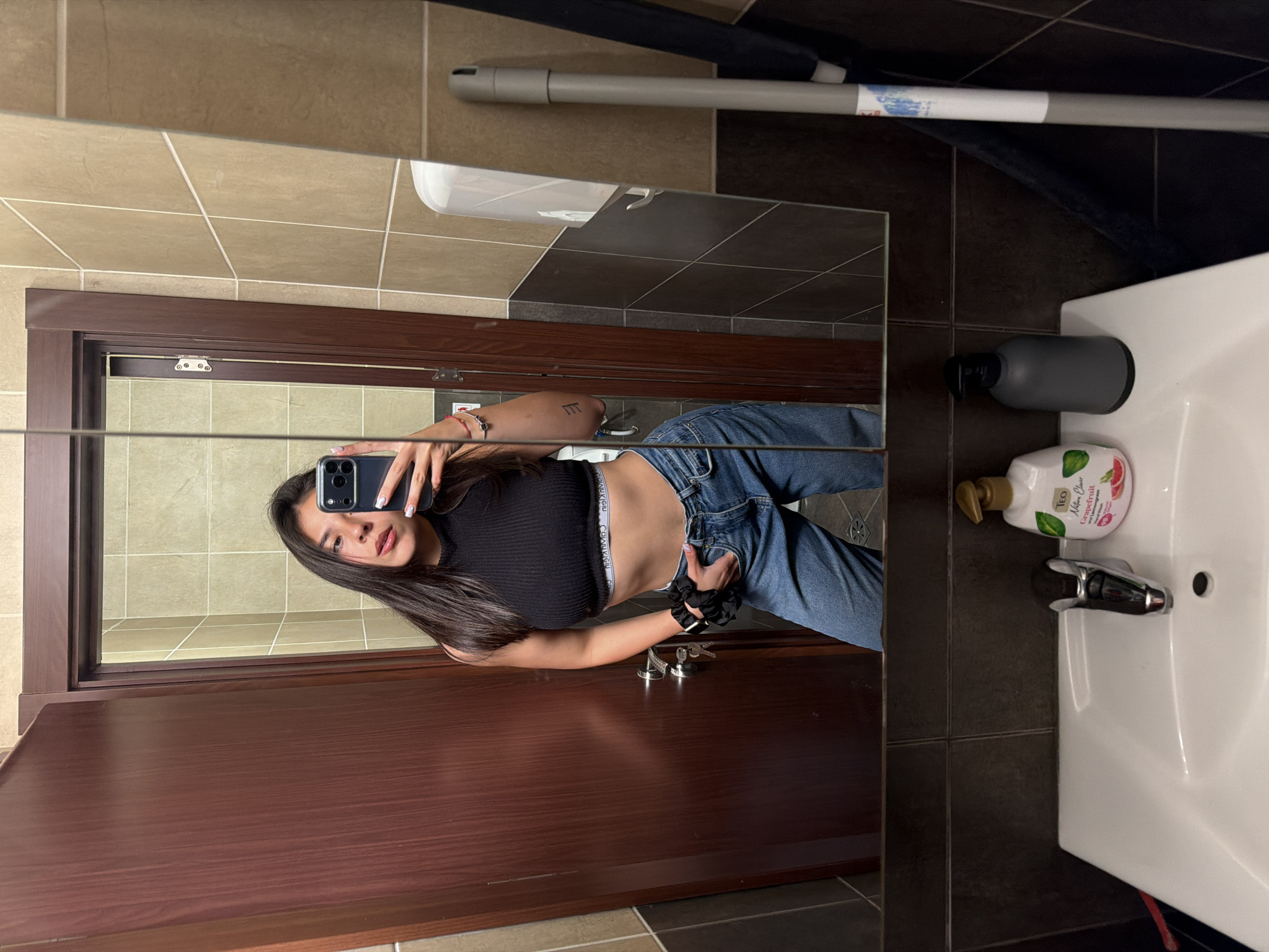A young woman is seen taking a mirror selfie in what appears to be a bathroom, located in Gabrovo, Bulgaria. She has long, dark hair and is dressed in a black ribbed crop top and blue jeans, revealing her midriff. On her left wrist, she wears a black scrunchie, and a small bracelet is visible on her right. Her right forearm displays a tattoo of three vertical lines. She holds a black smartphone, featuring multiple cameras, in her left hand to capture her reflection, while her right hand is positioned near the waistband of her jeans. The waistband of her underwear or undershirt, bearing the "Calvin Klein" brand name, is visible.
The bathroom features tiled walls with a mix of light beige/brown and darker brown/black tiles. A prominent dark brown wooden door with a frame and metal hardware is reflected behind the woman. To the right of the reflection, a white ceramic sink with a chrome faucet is visible, alongside a grey wall-mounted soap dispenser and a white bottle of "TEO Nature Clean Grapefruit" hand soap. In the upper section of the mirror's reflection, a long grey pole, possibly a cleaning tool, is seen. The floor is also tiled in a dark color. The setting indicates an indoor environment with artificial lighting.