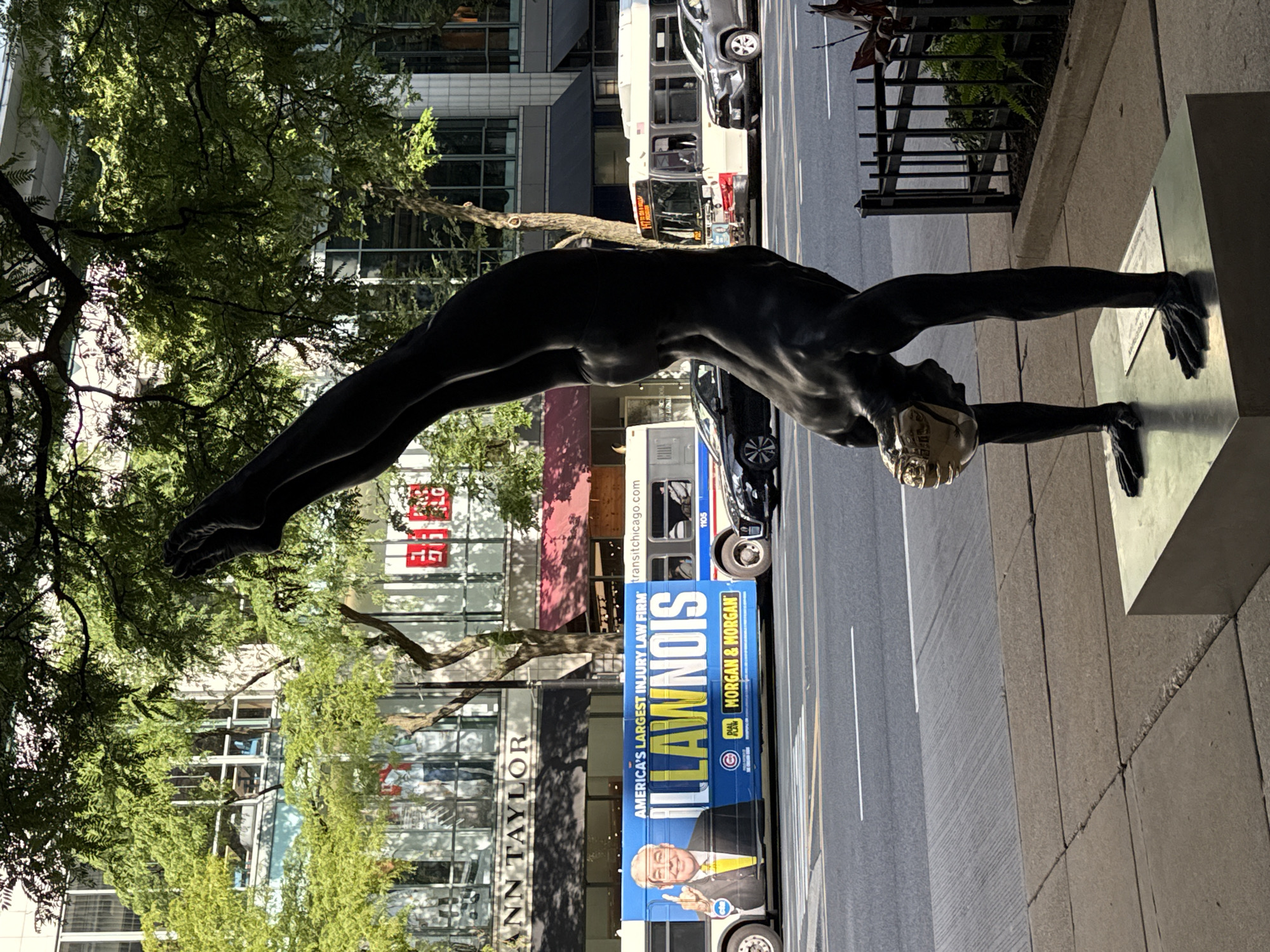 A statue of a dark-skinned gymnast in a handstand pose is positioned in the foreground on a city sidewalk, casting a shadow onto the sidewalk. The statue is on a metal pedestal, which has a plaque on it. Behind the statue, across a street, is a busy city street with vehicular traffic. A blue and white bus with a large advertisement for "ILLAWNOIS" lawyer firm "MORGAN & MORGAN" is prominently featured. The bus has a picture of a man in a suit pointing his finger. Another bus is partially visible behind it, along with several cars, including a black sedan and a gray SUV. Buildings line the street, with awnings and storefronts visible, including one for "ANN TAYLOR". Trees with green foliage partially obscure the view of the buildings and the sky. The scene appears to be in an urban setting, likely Chicago, given the "ILLAWNOIS" advertisement which plays on the state's name.