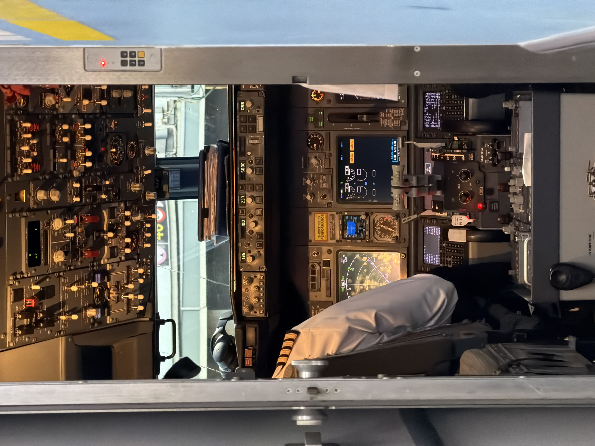 An interior view of an airplane cockpit in Palma de Mallorca, Spain, shows the complex array of controls and displays. A pilot, wearing a white shirt with gold stripes on the sleeve, is seated in the left pilot's chair, partially visible. The cockpit is filled with numerous buttons, switches, knobs, and multiple digital screens displaying flight information. Overhead, a panel of buttons and lights is visible. A stack of documents sits on a shelf above the main console. Headphones rest on a ledge above the displays. The lighting suggests it is daytime, with natural light filtering in from outside. The overall impression is one of advanced technology and focused operational readiness within the confined space of the aircraft's command center.