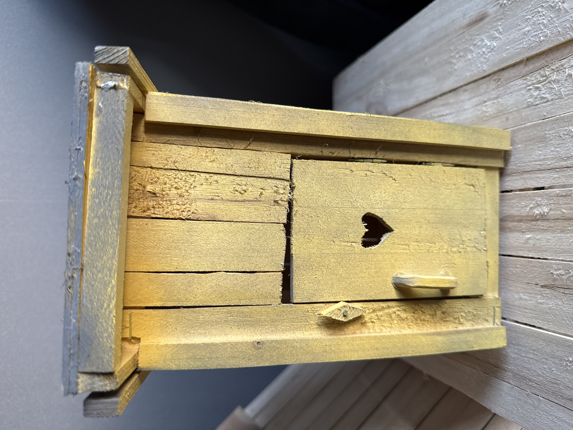 A close-up shot depicts a small, yellow wooden birdhouse. The birdhouse is constructed from small planks and features a heart-shaped opening. A small handle is affixed to the door. The birdhouse sits on a surface covered with pale wooden slats. The background is blurred, out of focus. There is no evidence of the location being Izhevsk, Russia.