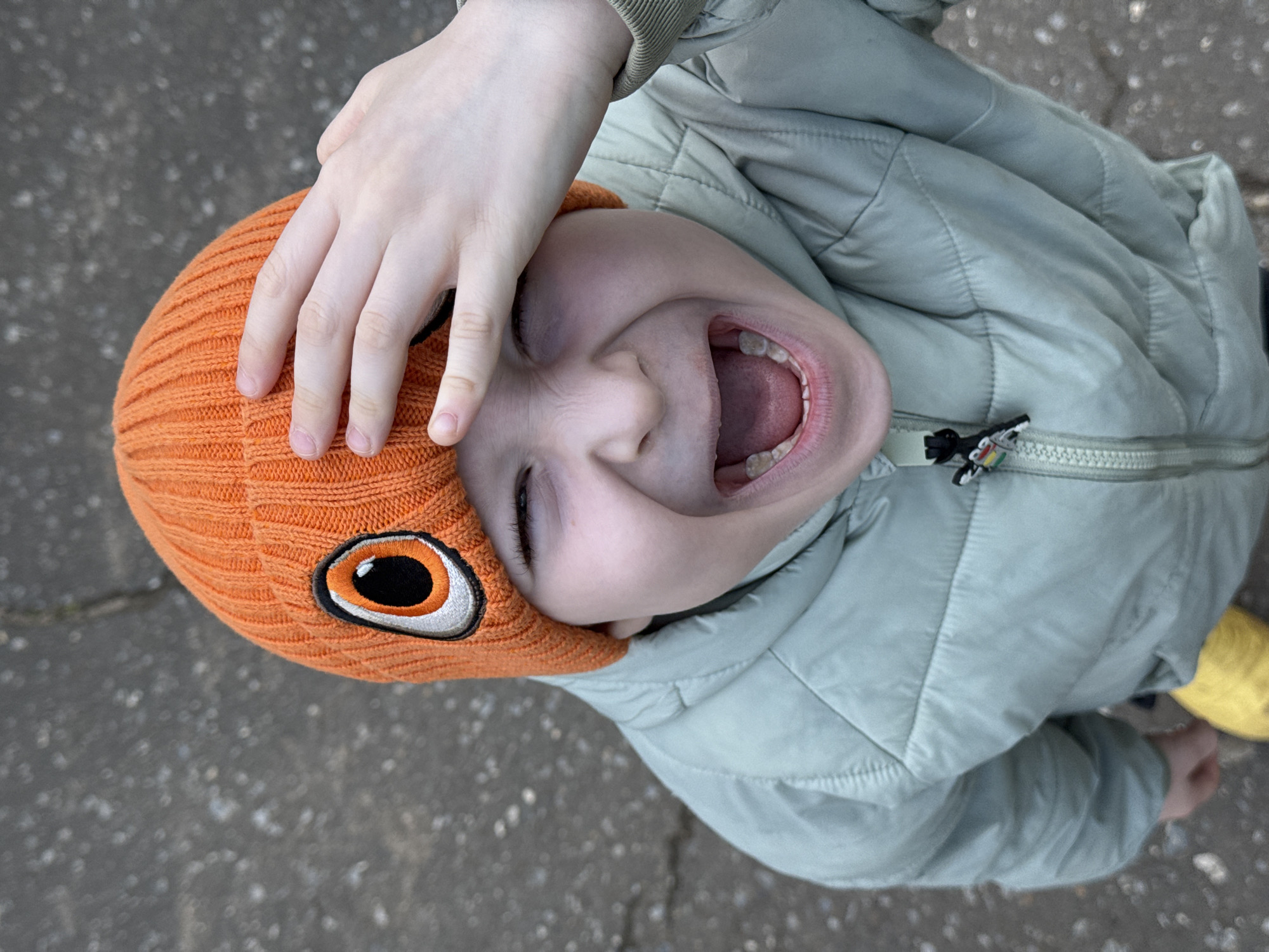 This is a top-down photograph of a young child, taken outdoors in what appears to be Chaikovsky, Russia. The child is looking directly up at the camera with their mouth wide open in a scream of joy or laughter, conveying strong emotions of excitement and playfulness. One hand is positioned on top of an orange knitted beanie, which covers the child's head. The beanie features a prominent, embroidered cartoonish eye design on its side. The child is wearing a light green padded jacket, which is partially visible, showing its zippered front and a small, colorful zipper pull near the collar. The background is a blurry, mottled grey surface, consistent with asphalt or concrete pavement, indicating an outdoor urban or suburban setting. The natural lighting is soft, suggesting an overcast day or perhaps early morning/late afternoon. No visible text is present in the image. The overall impression is one of high energy and cheerful exuberance.