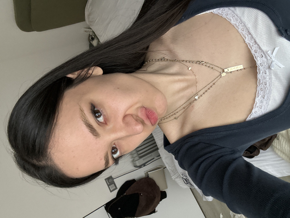 A young female, appearing to be in her late teens or early twenties, is depicted in a close-up selfie, occupying the majority of the frame. Her dark, long, straight hair is parted slightly off-center and extends over her shoulders. She possesses brown eyes and light brown eyebrows, with minimal visible makeup. Her mouth is pursed in a pouting expression, and her gaze is directed towards the camera. She is wearing a dark blue, long-sleeved top, possibly a cardigan or shrug, over a white camisole with white lace trim along the neckline and a small white bow at the front center. Multiple gold-colored necklaces are visible; the longest features a rectangular pendant inscribed with "MARI" and additional text above it. The background indicates an indoor setting, likely a residential room with light-colored walls. To the left, a wall-mounted light switch or electrical outlet is visible above a vertical radiator. Below the radiator, a dark brown, possibly suede, bag with a lighter strap hangs on the wall. To the right, part of a bed with a white comforter and an olive-green headboard is visible. The image contains no explicit visual markers to confirm the location as Yerevan, Armenia.