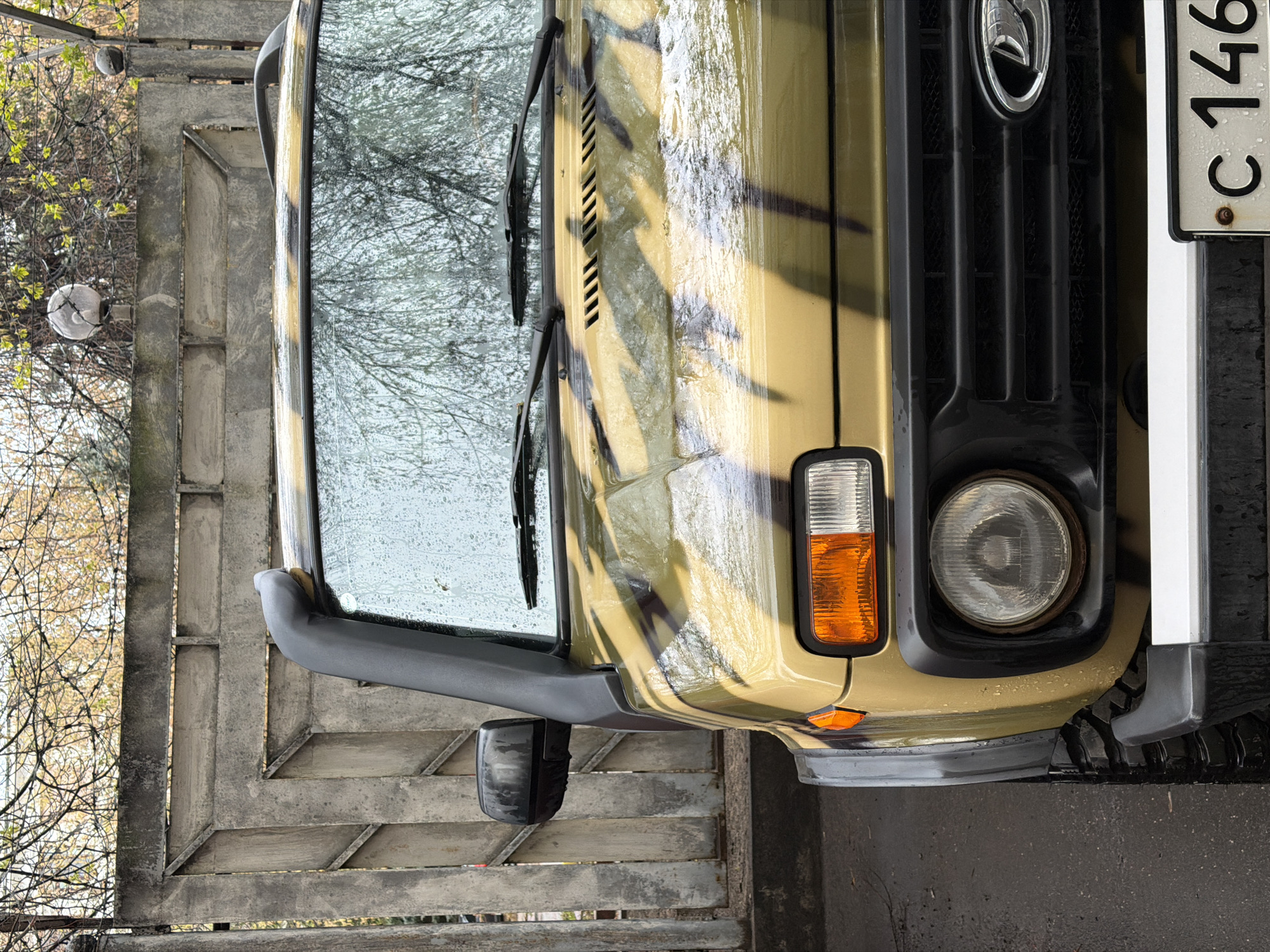 The image shows the front of a Lada Niva car in a camouflage pattern, parked on a concrete surface. The windshield is wet with rain. The license plate is partially visible, showing the letters "C" and the numbers "146". The background includes a grey metallic fence, trees, and two globe-shaped light fixtures. It's a rainy day and the picture was taken in Moscow, Russia.