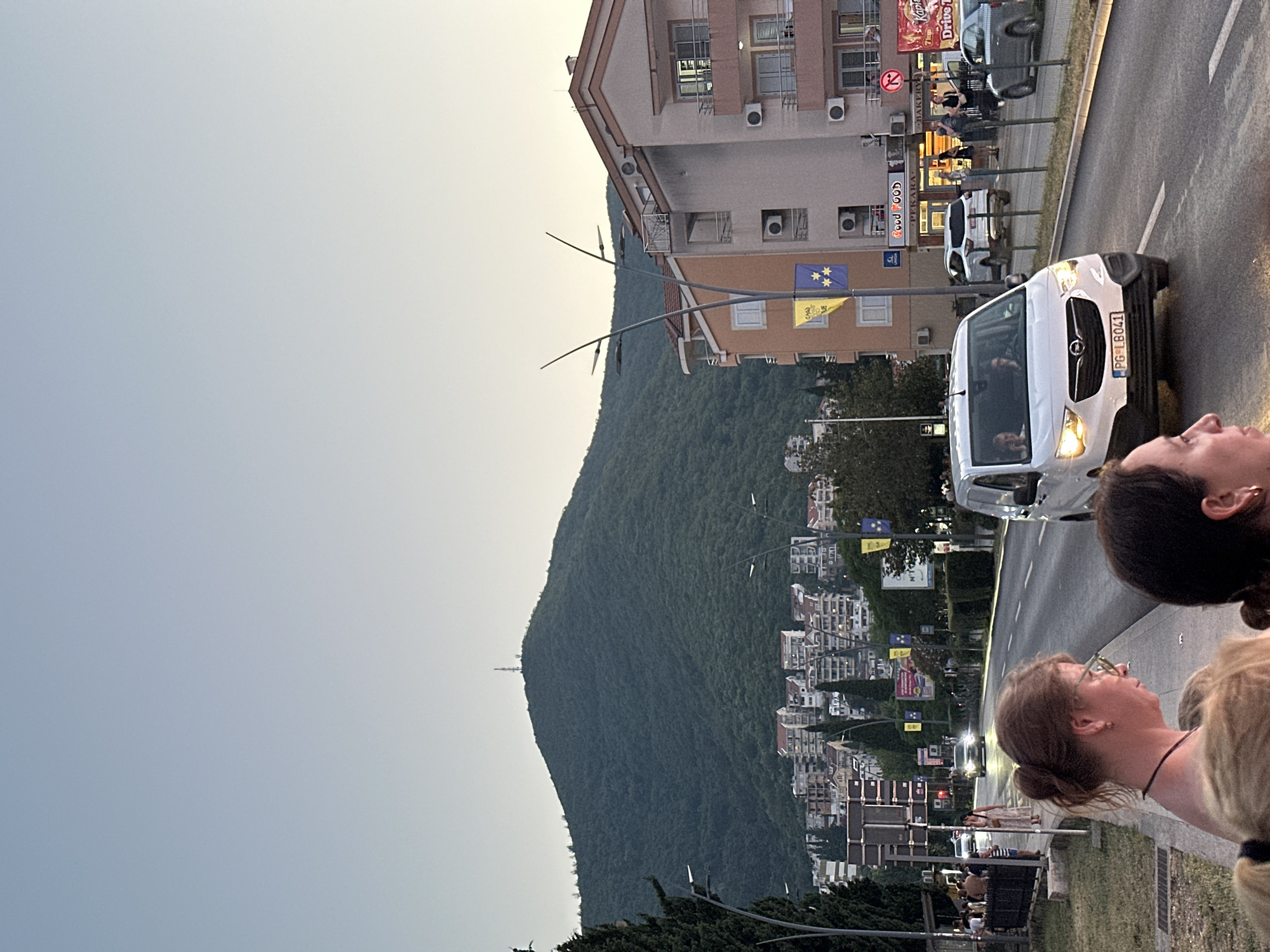 The image captures a street scene in Budva, Montenegro, likely during the evening or early morning given the soft, diffused light and clear sky. In the foreground, two women are partially visible, facing away from the camera and looking towards a street. One woman, with glasses and her hair in a bun, stands on a sidewalk.
The street is lined with buildings and a lush, tree-covered mountain rises in the background, topped with a communication tower. Cars are present on the road, including a white van with its headlights on, and a white SUV. A row of buildings with balconies and air conditioning units are situated along the street. Signs on the buildings indicate businesses such as a bakery. Streetlights with multiple lamps illuminate the area. The sidewalk appears to be made of concrete, and there is a grassy area to the left. People are visible on the sidewalk, some walking and others appearing to be waiting. The overall impression is of a bustling urban area adjacent to a natural landscape.