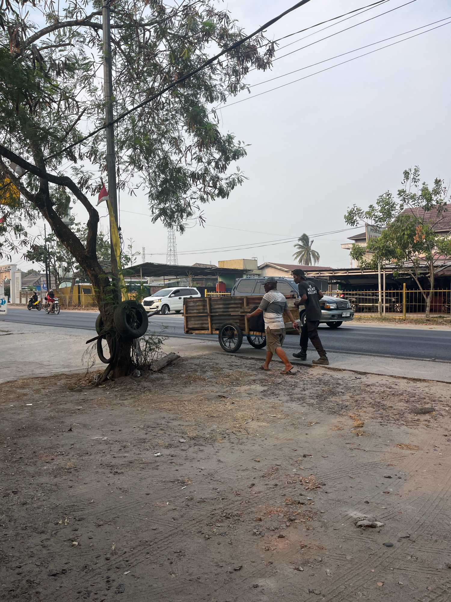 In the city of Kayu Agung, Indonesia, two men are seen pushing a wooden cart with wheels along a dirt path next to a paved road. The cart appears to be loaded with something, though it is not clearly visible. The man in front is wearing a striped shirt and shorts, while the man behind him is wearing a dark t-shirt and pants. They are both walking towards the right side of the frame.
The environment is a street scene with a tree on the left side of the image. The tree has several old tires wrapped around its trunk. Power lines are visible overhead, and a utility pole is situated near the tree. In the background, there are buildings and other vehicles on the road, including a white SUV and a dark sedan. The sky is overcast, suggesting it might be late afternoon or early morning. The overall scene depicts a common street activity in a developing urban area, with elements of both traditional transport and modern infrastructure.