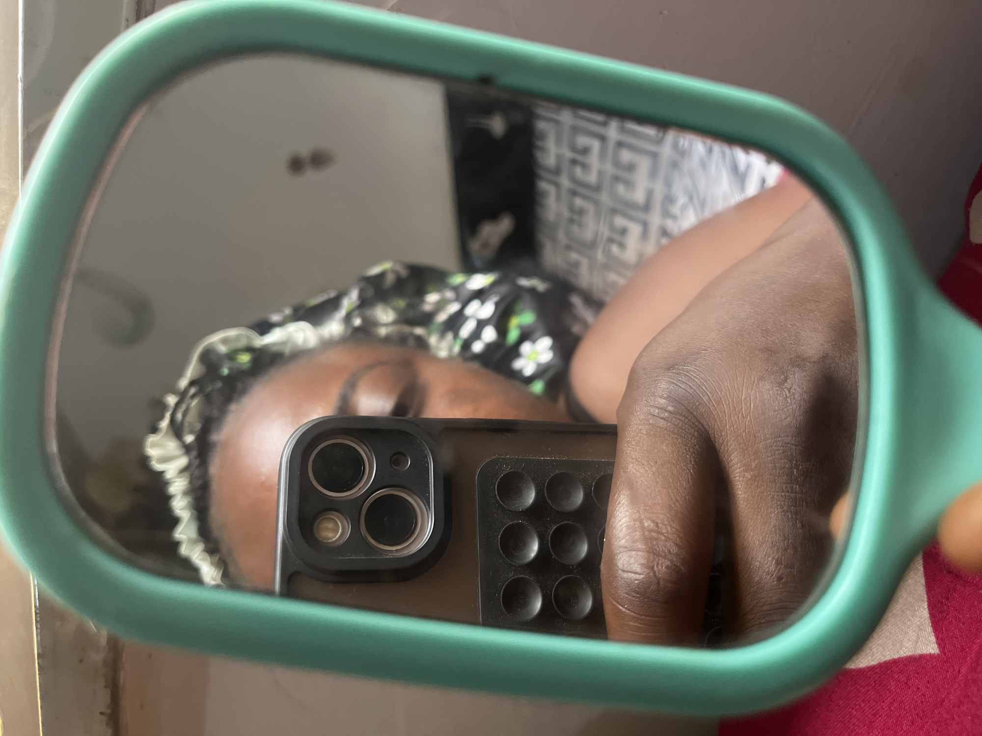 The image is a selfie taken in a mirror. The person holding the phone is in the foreground, obscuring part of the mirror's reflection. The person in the mirror appears to be lying down, wearing a head covering with a floral pattern. The background is blurry and difficult to discern, but it appears to show a wall with a patterned design and potentially a red fabric. It is not possible to determine the location as being in Abuja, Nigeria from the given information.