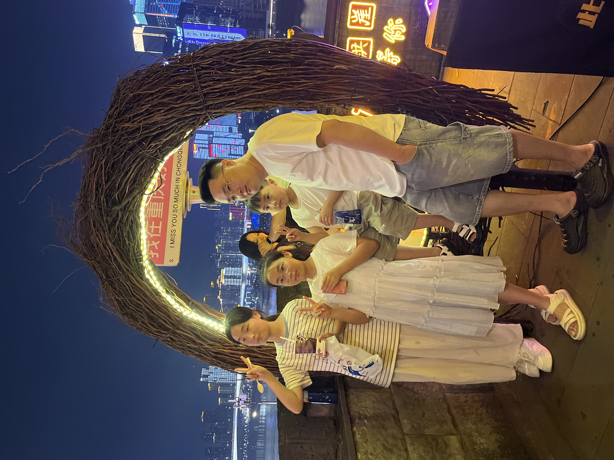 In Yuzhong District, China, a family of four poses for a photo under a large, twig-like arch adorned with yellow lights. The arch frames a nighttime cityscape with illuminated buildings and a body of water. The family includes a man in a white t-shirt and denim shorts, a woman in a white dress, and two children, a boy and a girl, both wearing t-shirts. The two girls are making peace signs with their fingers. The man has his hand in his pocket. The background shows a vibrant urban landscape at night.