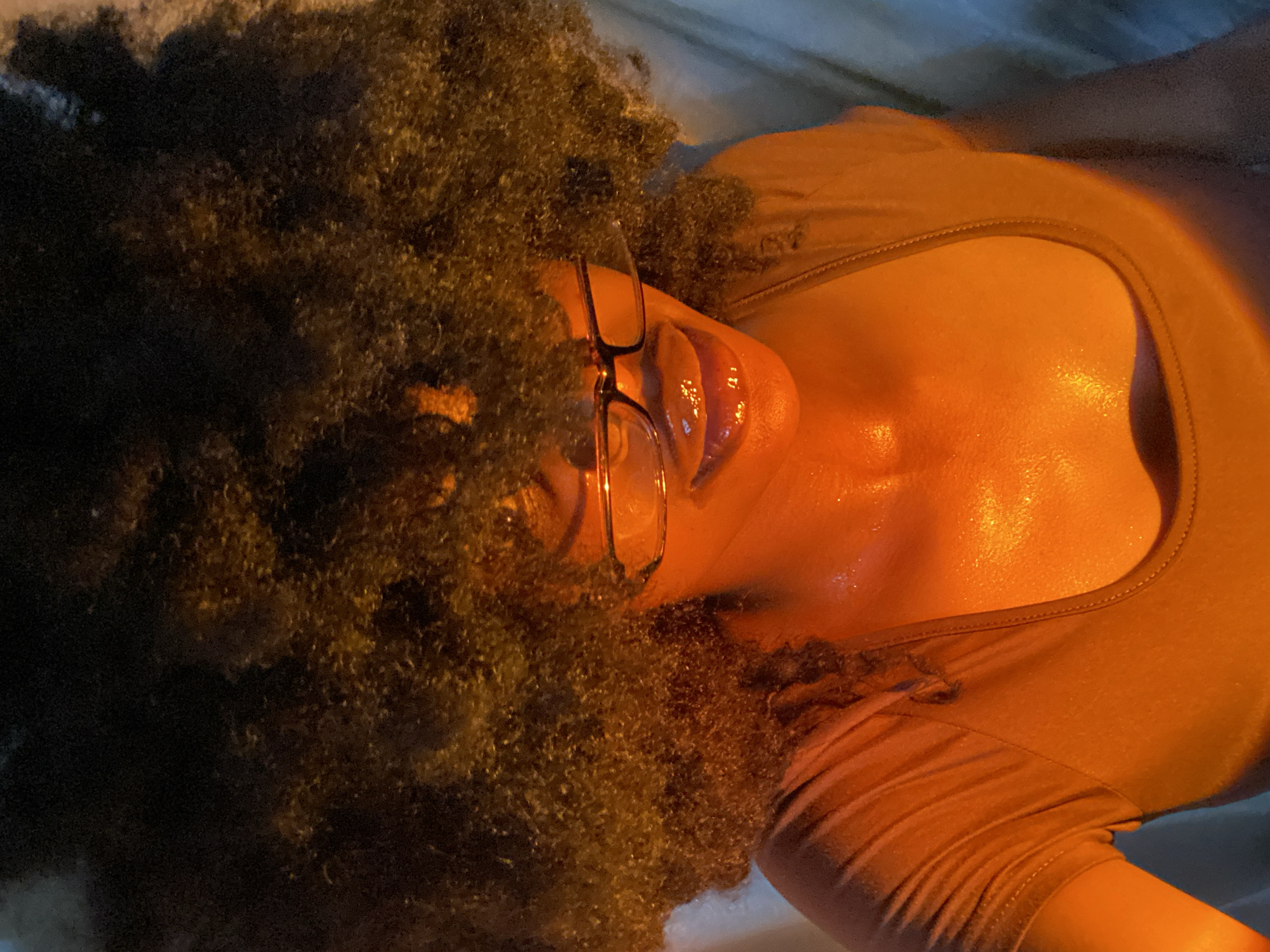A woman with an afro and glasses is wearing a brown top. The photo is taken at an angle, and her face and chest are illuminated, appearing wet or shiny. The background is unclear. It is not possible to identify the location in Iranla, Nigeria from this image.