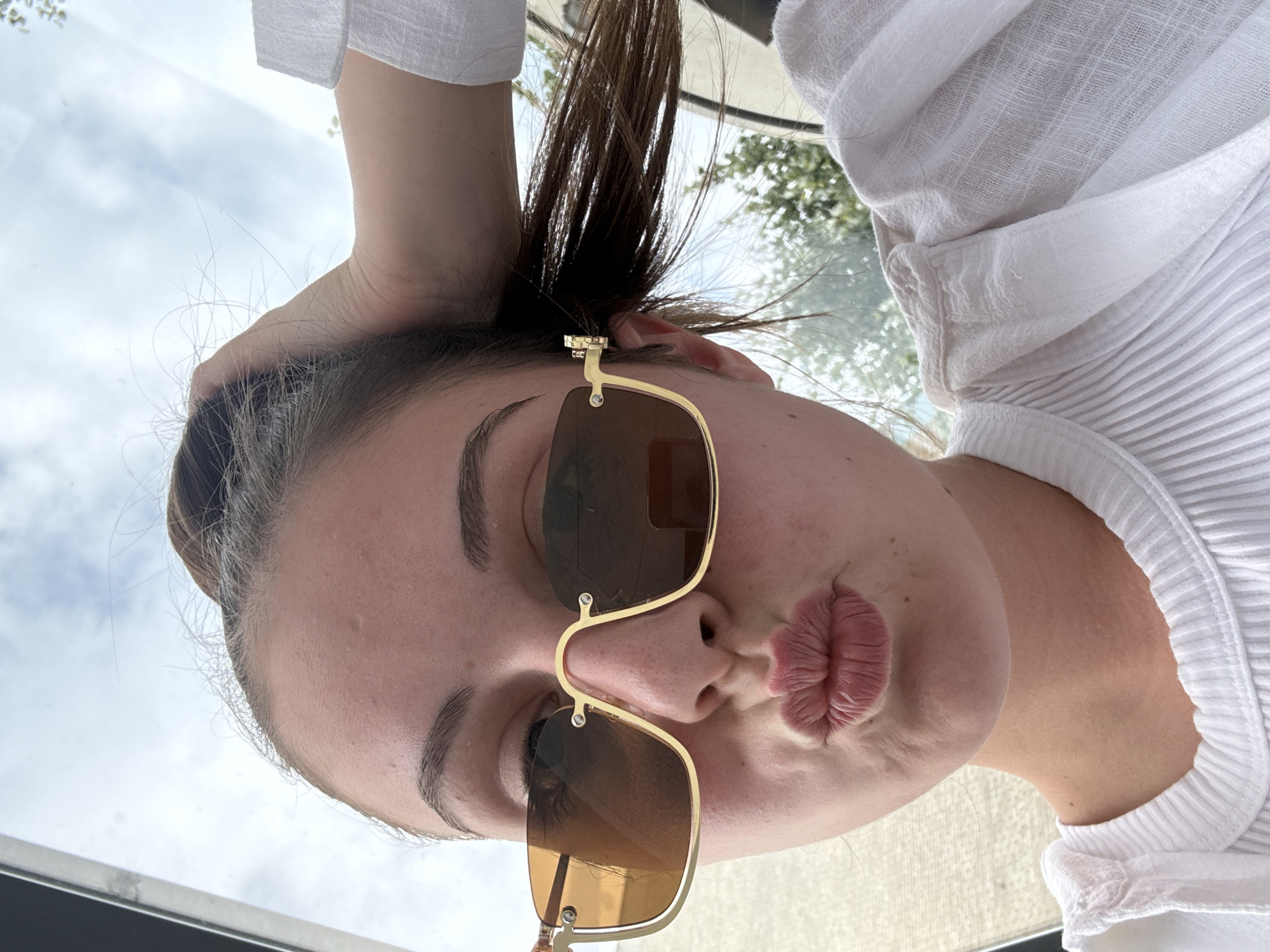 A young woman with brown hair pulled back is captured in a close-up, low-angle selfie. She is wearing oversized, gold-rimmed sunglasses with amber lenses and a white ribbed shirt. Her lips are pursed as if for a kiss. Her right hand is raised, touching her hair. The background consists of a cloudy sky and some blurred greenery, suggesting an outdoor setting. The image appears to have been taken on a sunny day. The specific location in Burgas, Bulgaria, is not discernible from the image itself.